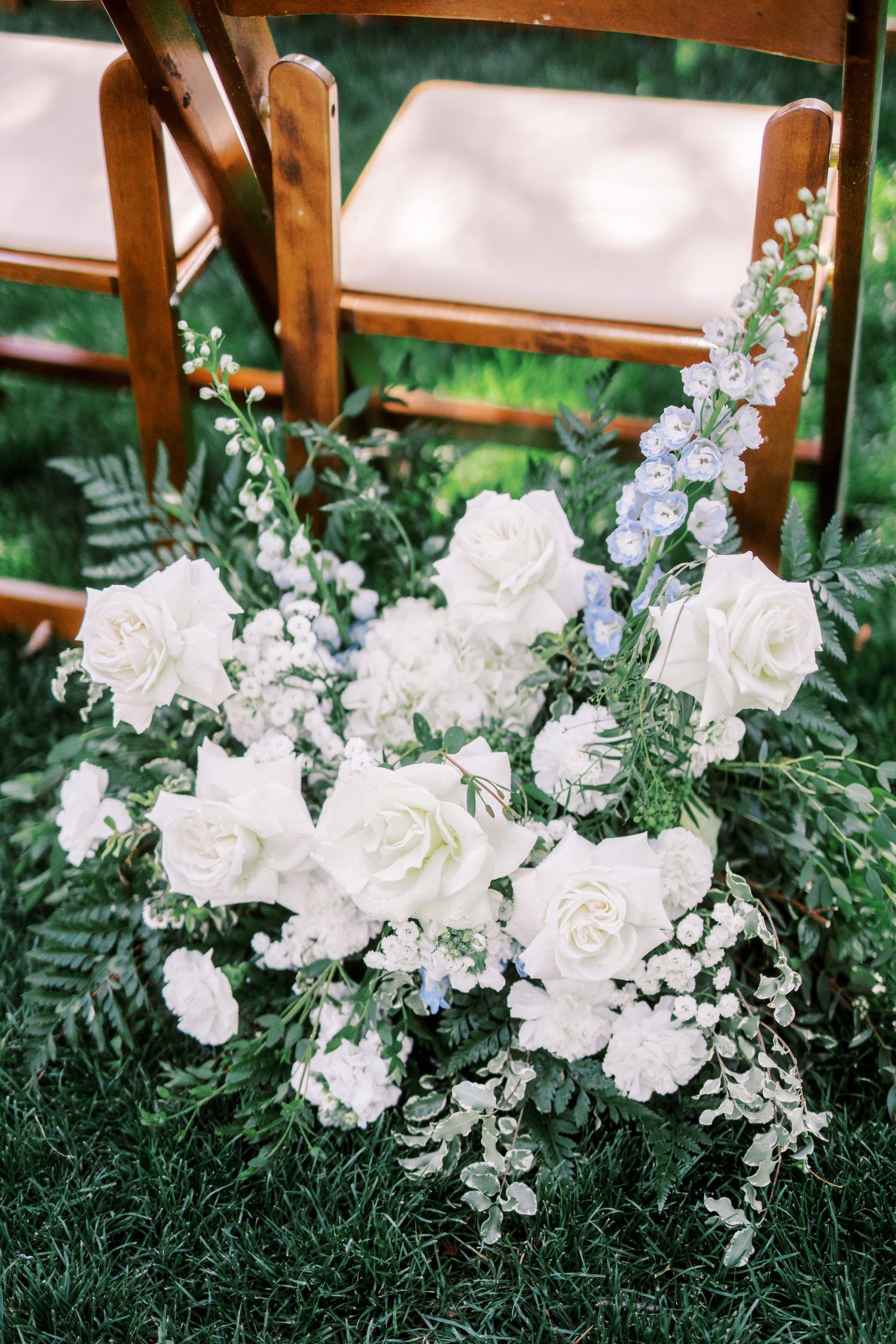 Beautiful white rose and delphinium floral arrangement with ferns sits on grass near wooden chairs at an outdoor wedding.