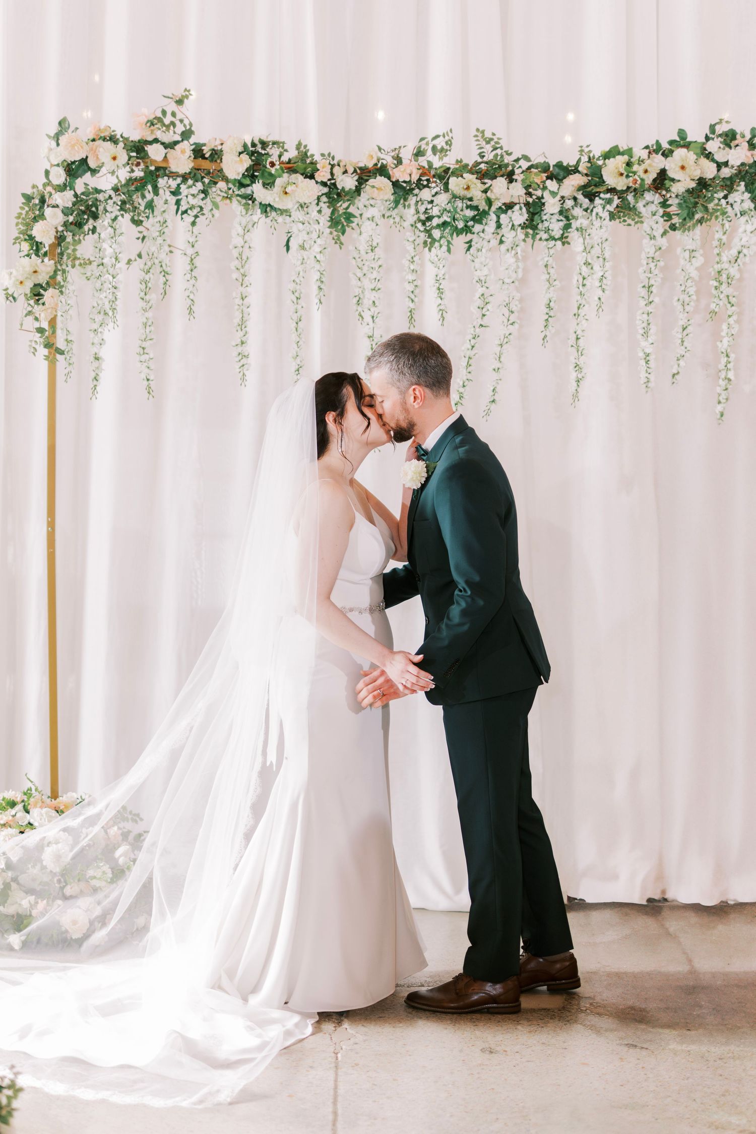 Wedding couple embraces under an elegant floral arch with cascading white flowers and greenery.