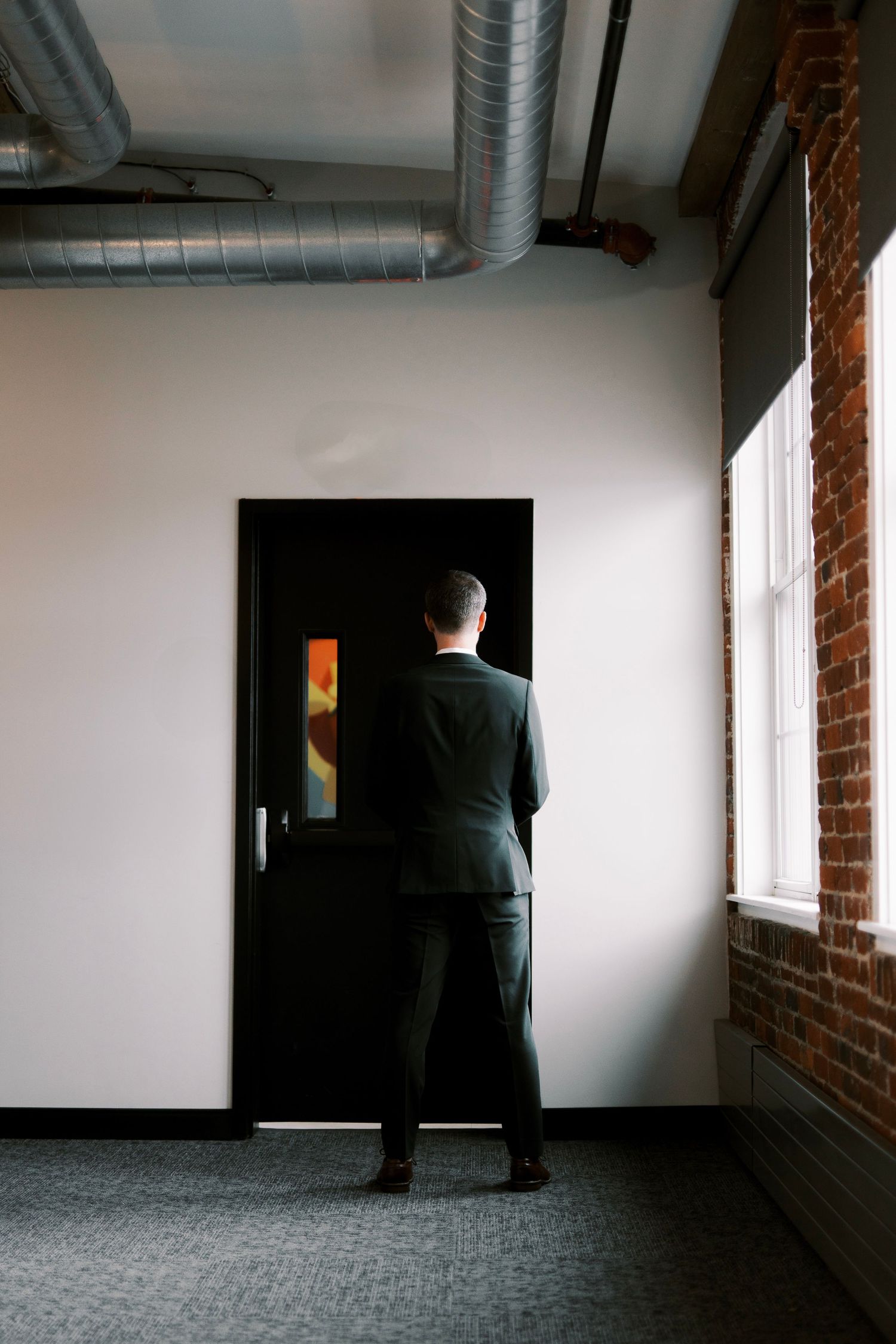 A person in a dark suit stands before a black door in an industrial hallway with exposed ductwork and brick walls.