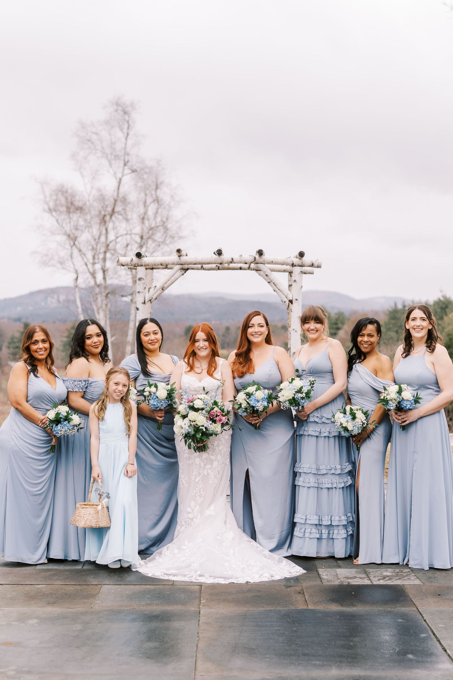 A wedding party wearing elegant powder blue bridesmaid dresses poses outdoors under an archway.