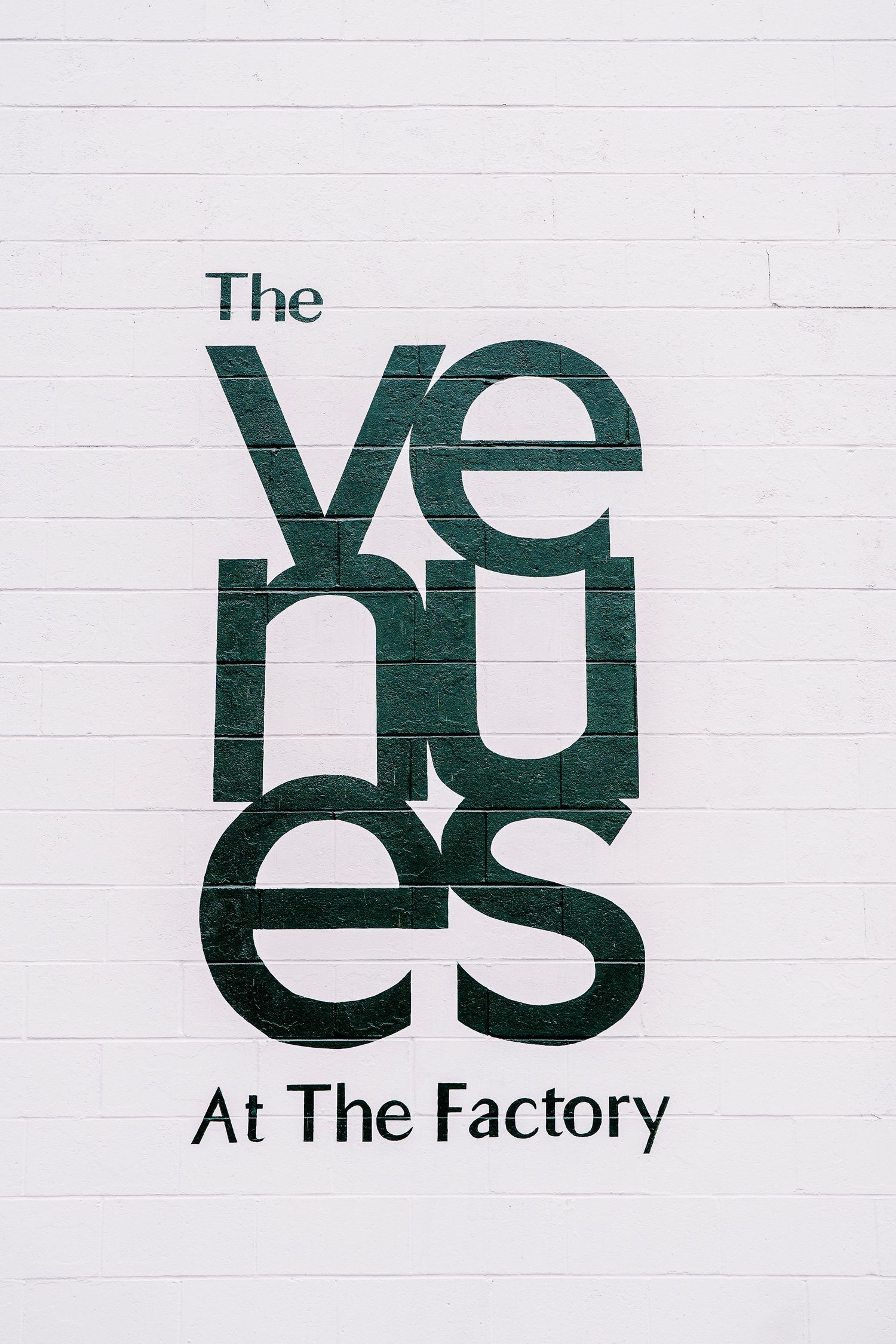 The Venues at The Factory logo in dark green text on white painted brick wall.