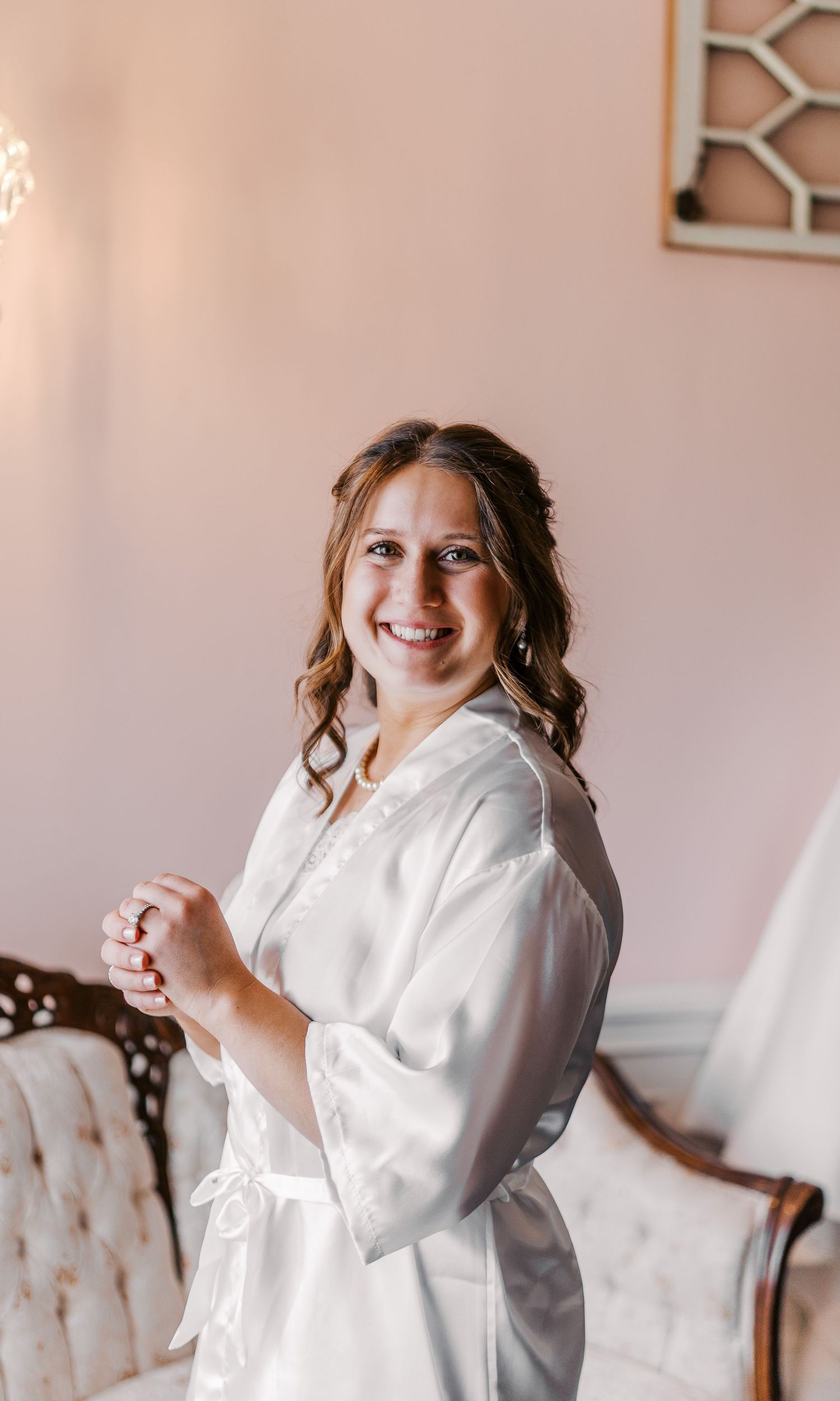A joyful figure wearing a white robe poses playfully against a pink wall beside a decorative chair.