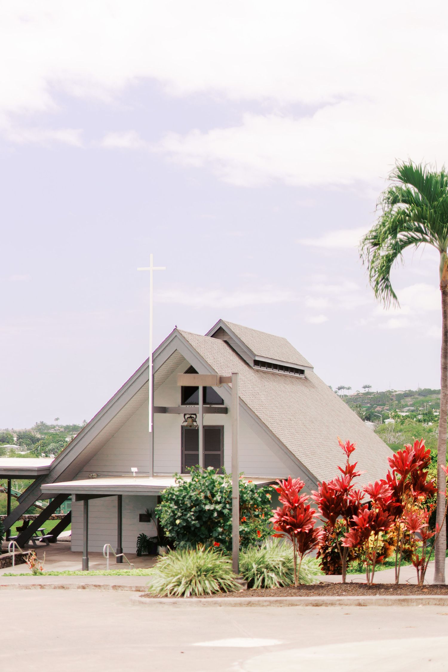 Modern A-frame church with white exterior and tropical landscaping featuring red flowering plants and palm trees.