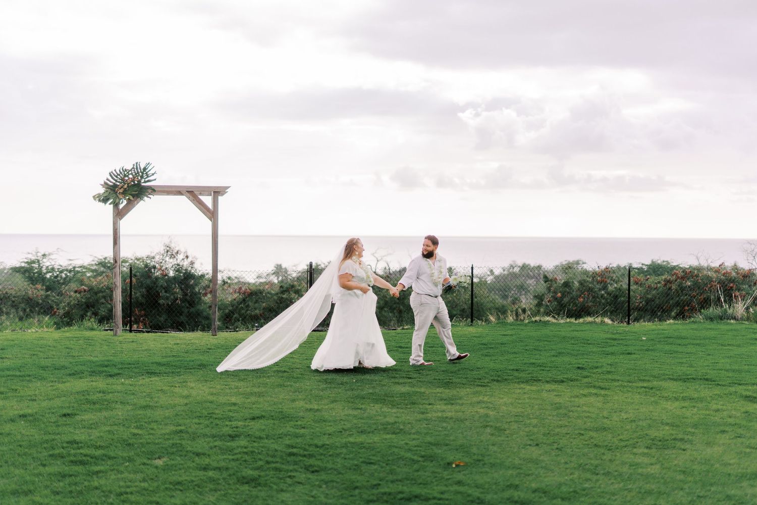 Couple in wedding attire standing on green lawn with wooden arch in background.
