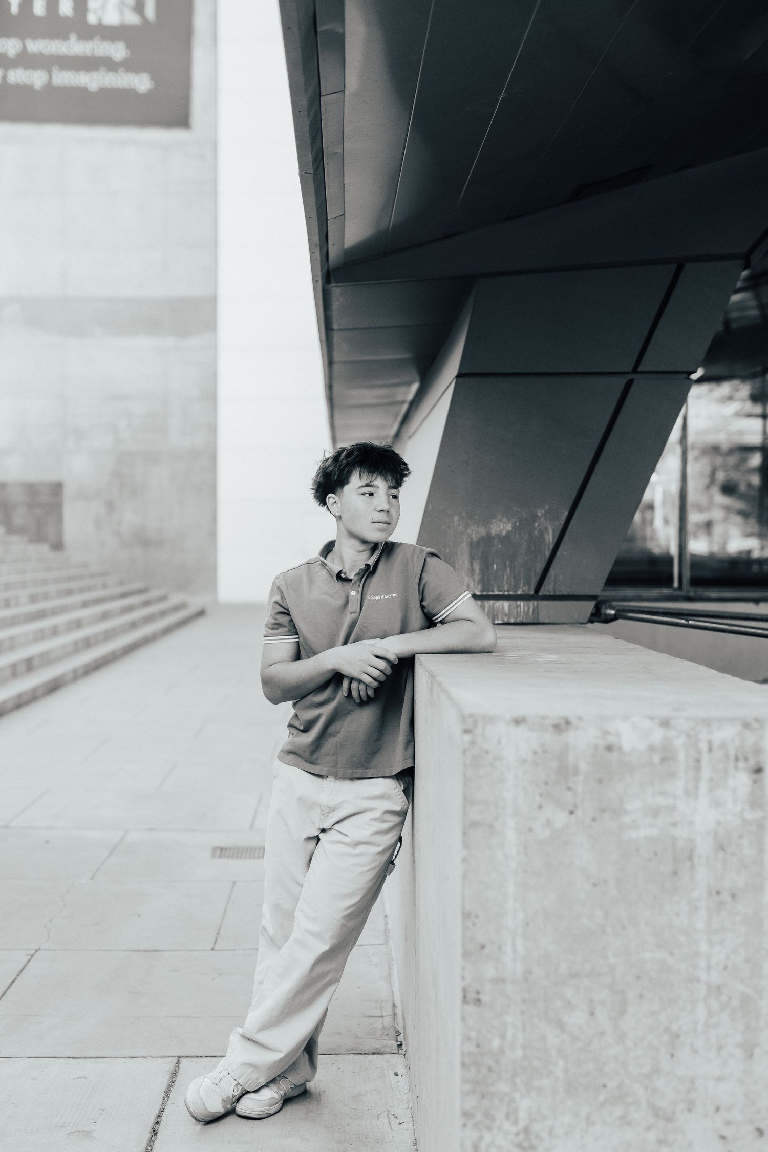 Young person in casual attire leaning against a modern architectural wall in a black and white photo.