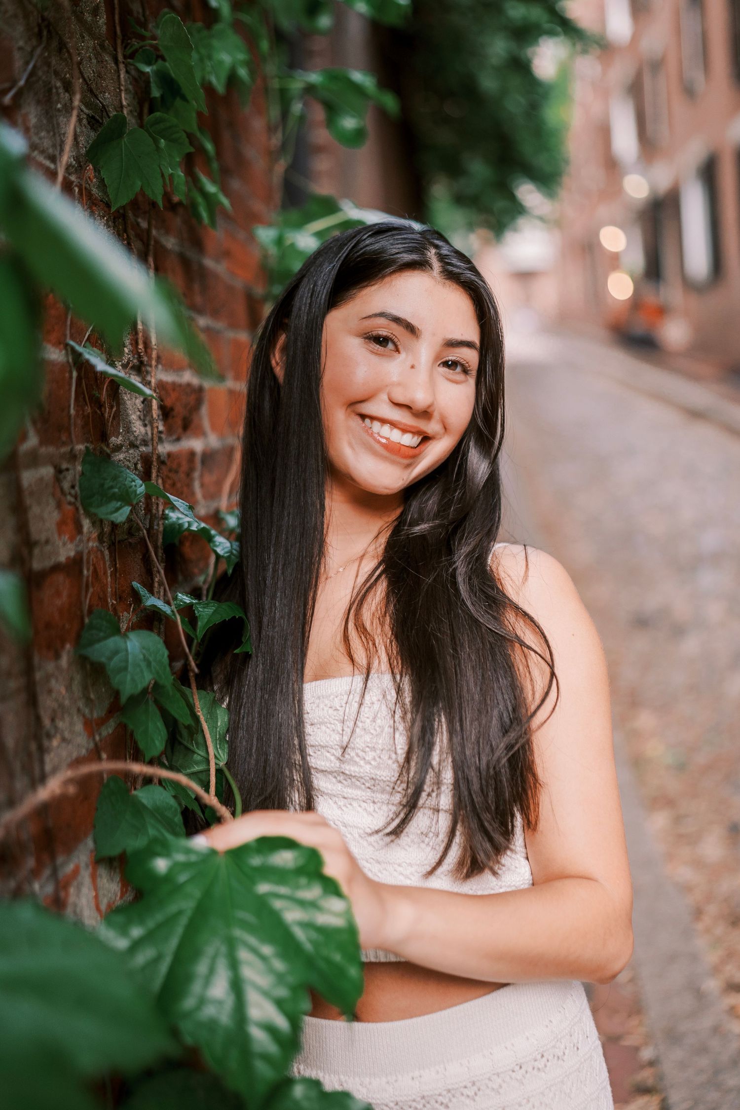 A smiling woman in white lace dress poses against a brick wall with ivy on a charming cobblestone street.