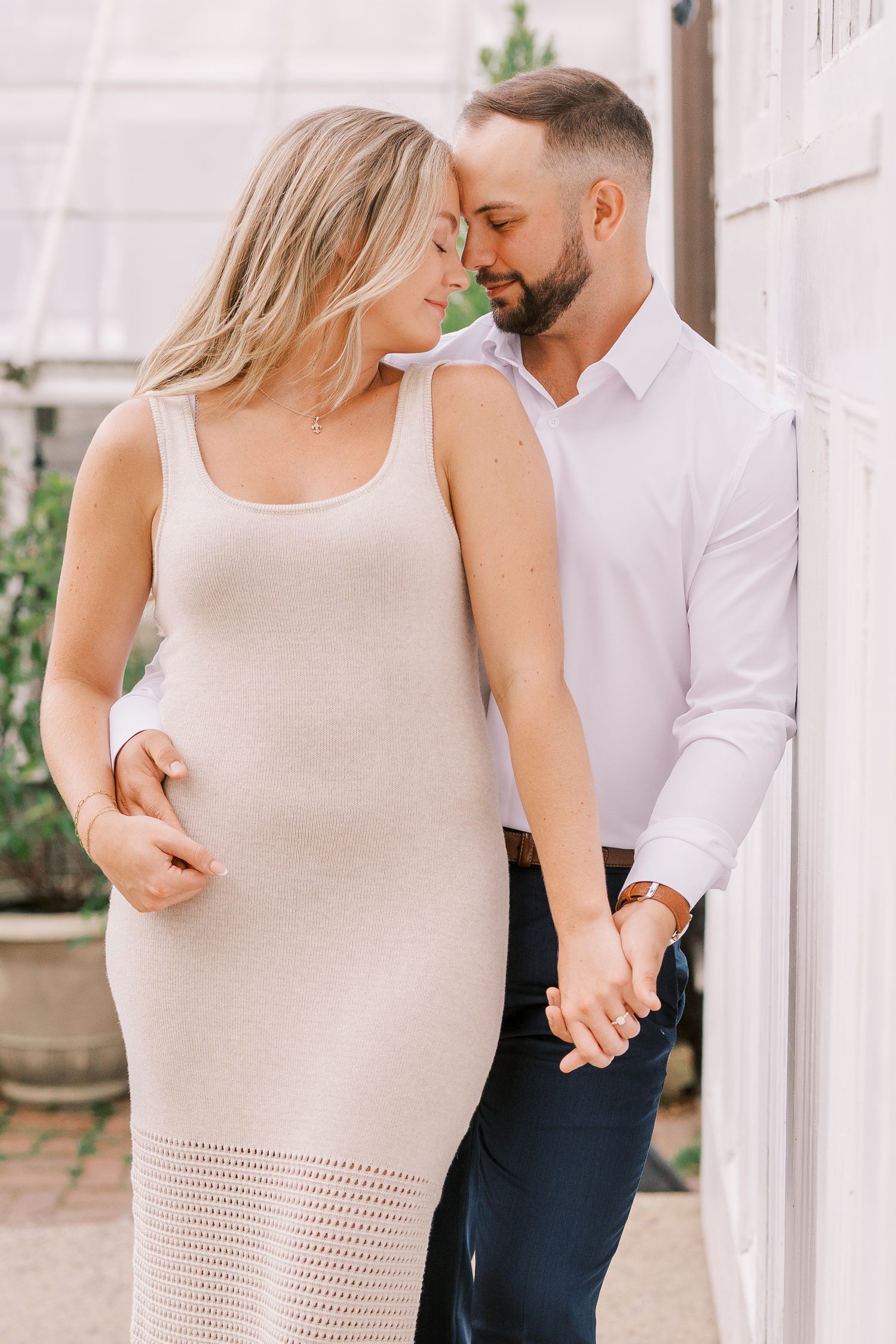 Intimate outdoor portrait of couple in light-colored clothing holding hands and leaning close together against white wall.