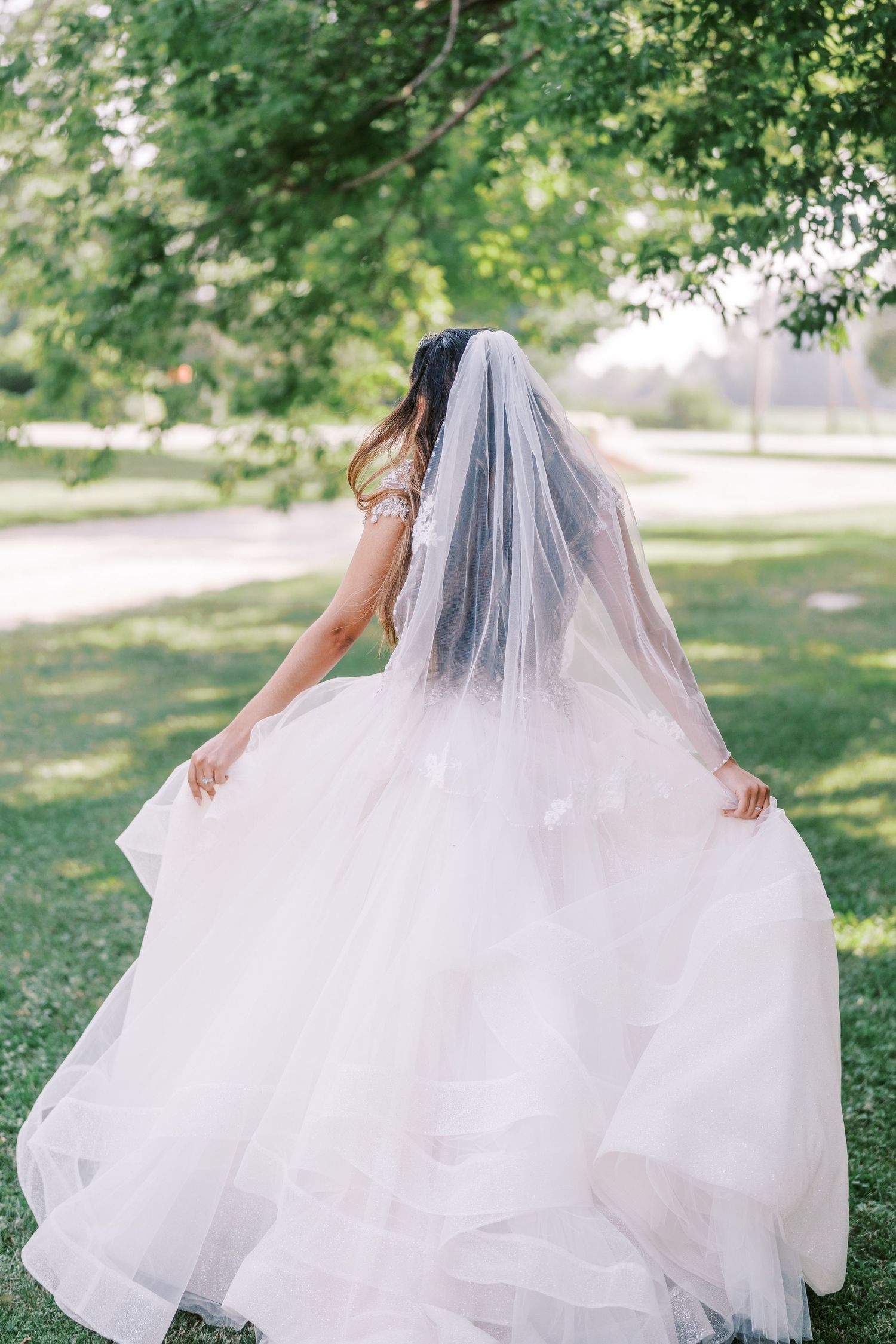 A bride in a flowing white tulle gown and veil spins gracefully in a sunlit garden setting on her wedding day.