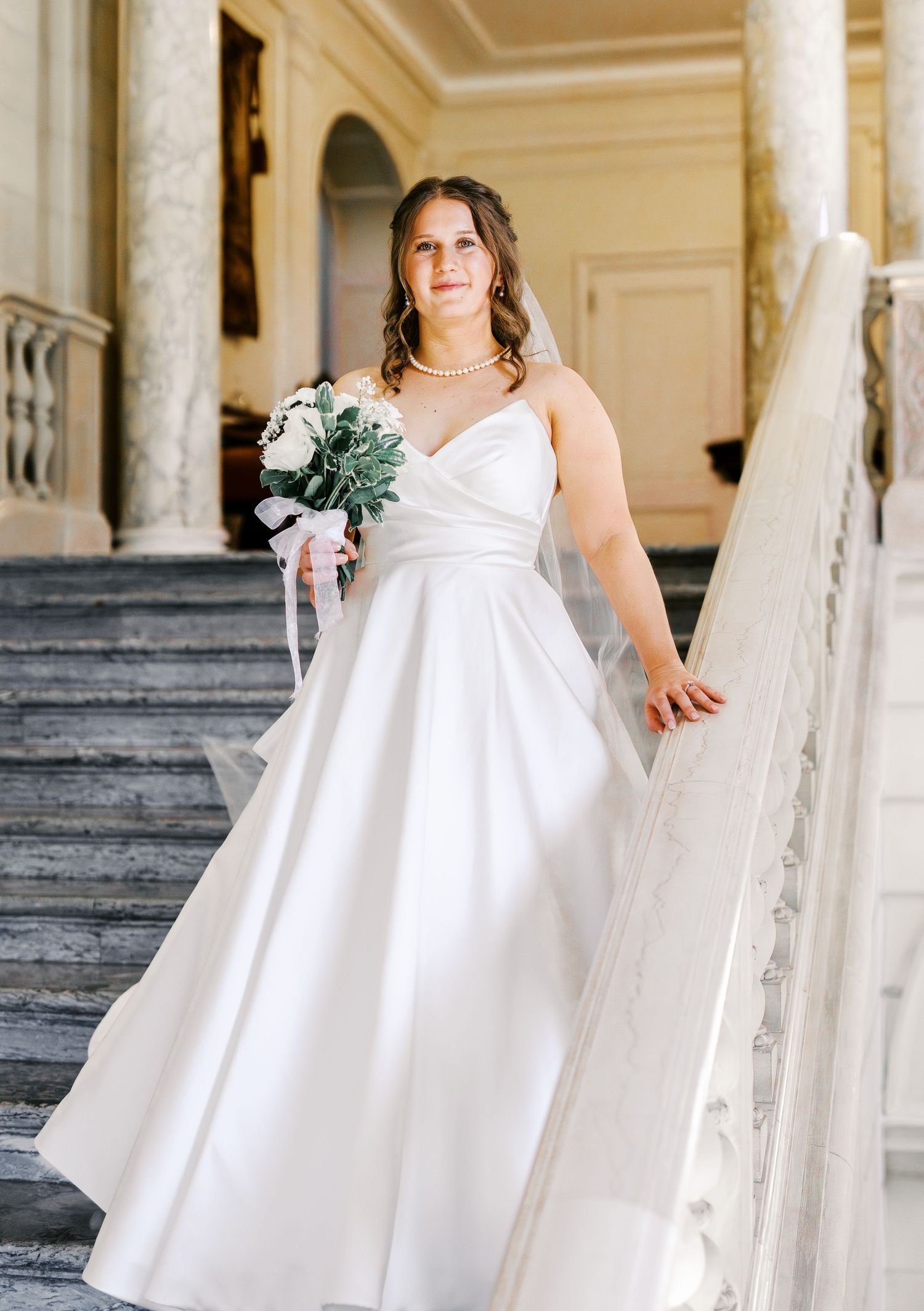 Bride holding bouquet poses on grand staircase in classic white A-line wedding dress.