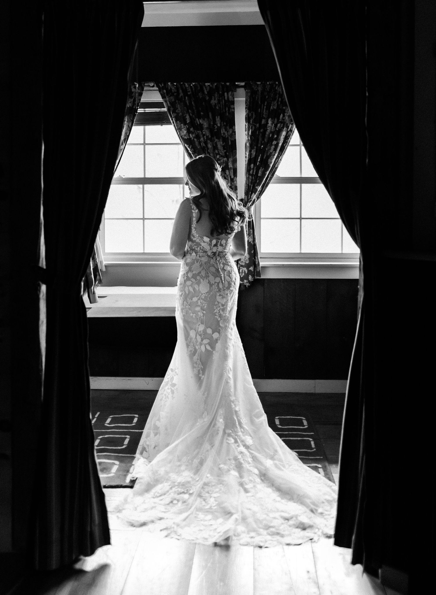 A dramatic black and white silhouette of a bride in a lace gown with long train by a window.