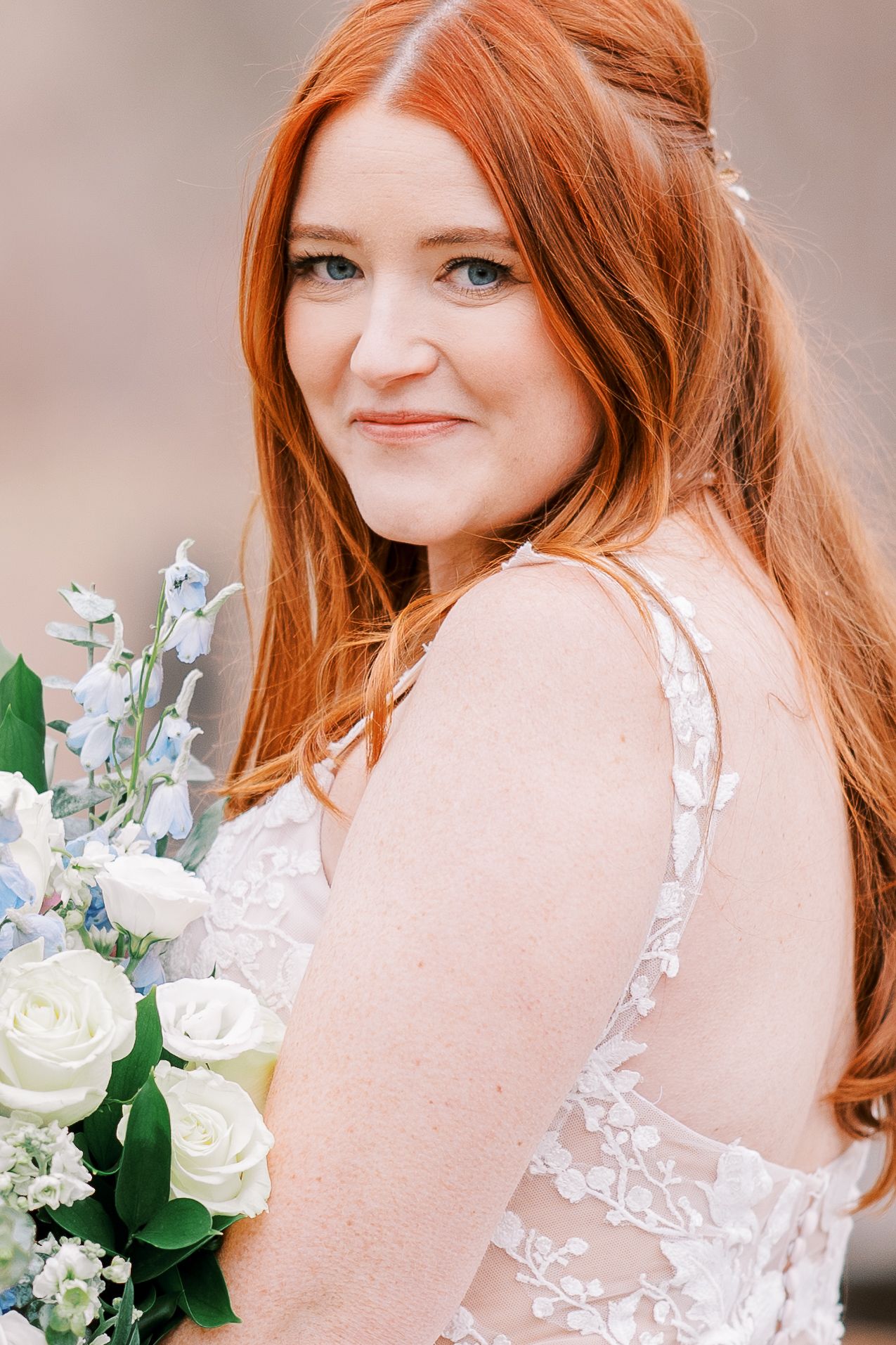 A bride in a lace wedding dress holds a bouquet of white roses and delicate flowers while smiling over her shoulder.