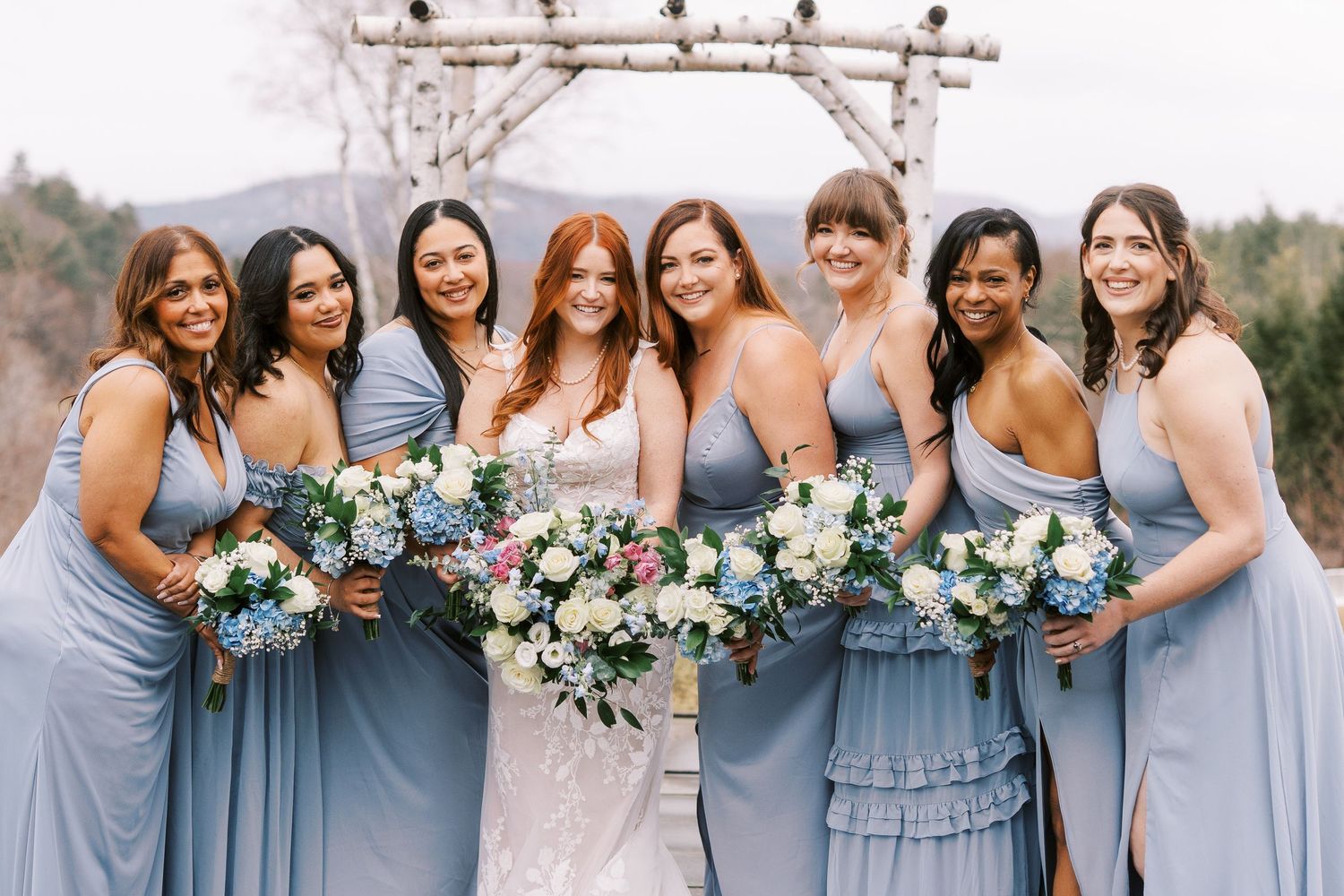 A wedding party lineup wearing dusty blue bridesmaid dresses holds elegant white and blue floral bouquets.