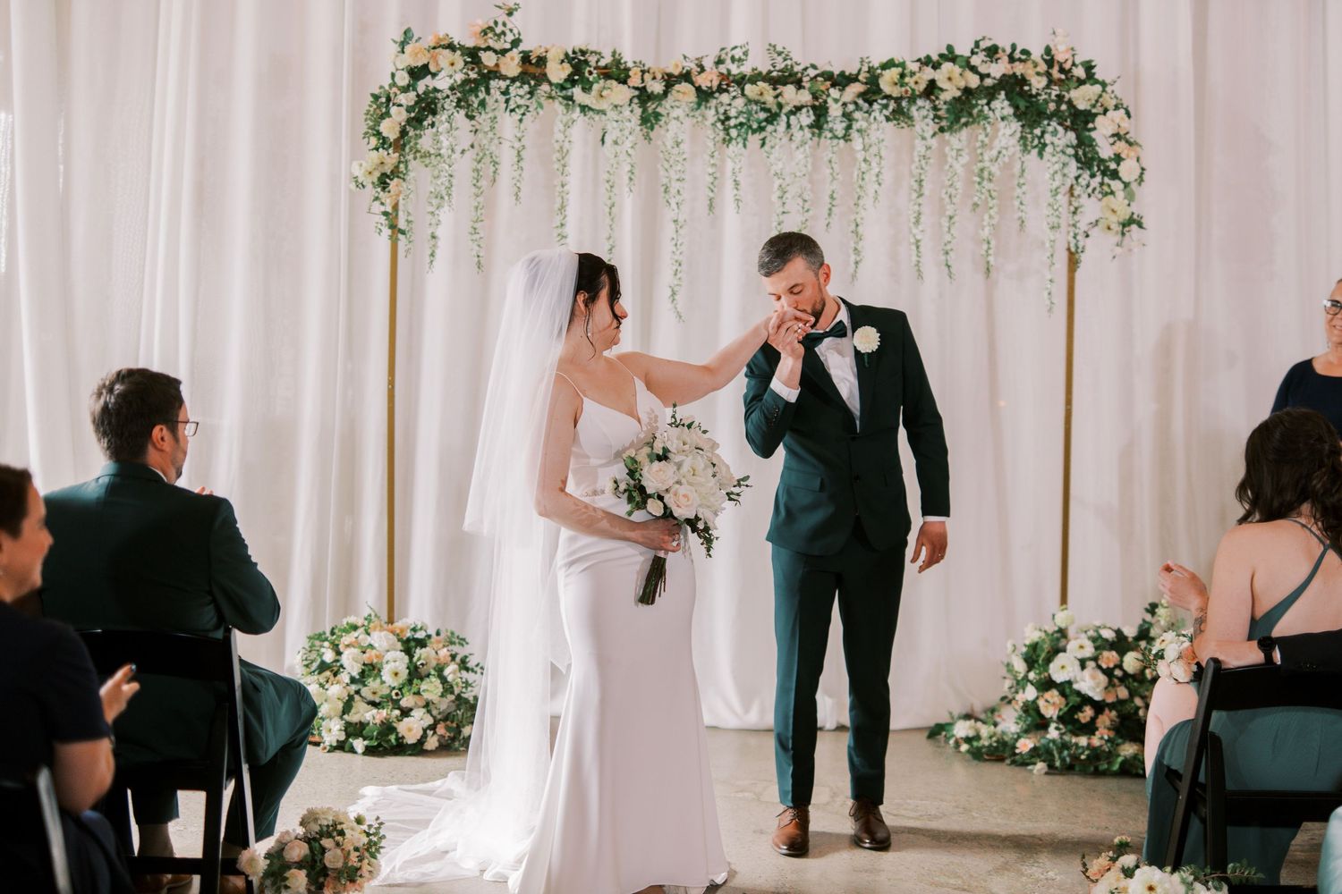 A couple shares a sweet moment during their wedding ceremony under a floral arch with white curtains and greenery.