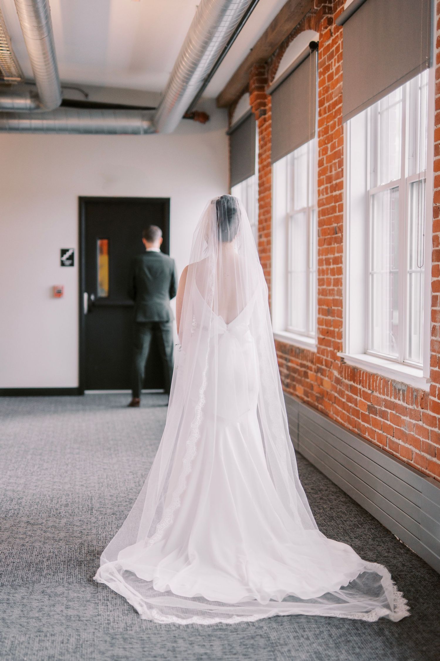 A flowing white wedding veil trails behind as a figure stands in a modern hallway with brick walls.