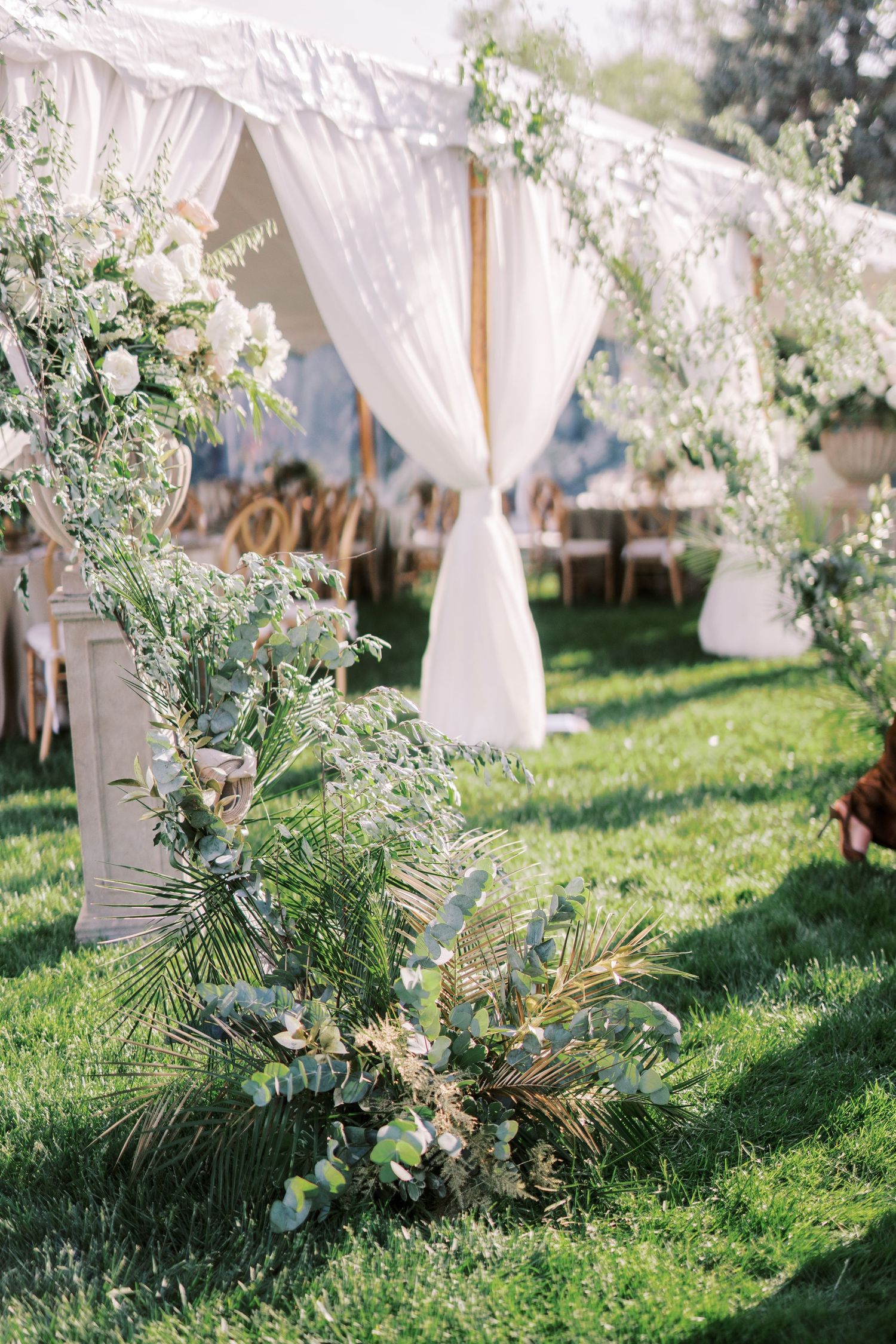 Elegant outdoor wedding tent with flowing white drapes and lush greenery floral arrangements on a sunny grass lawn.