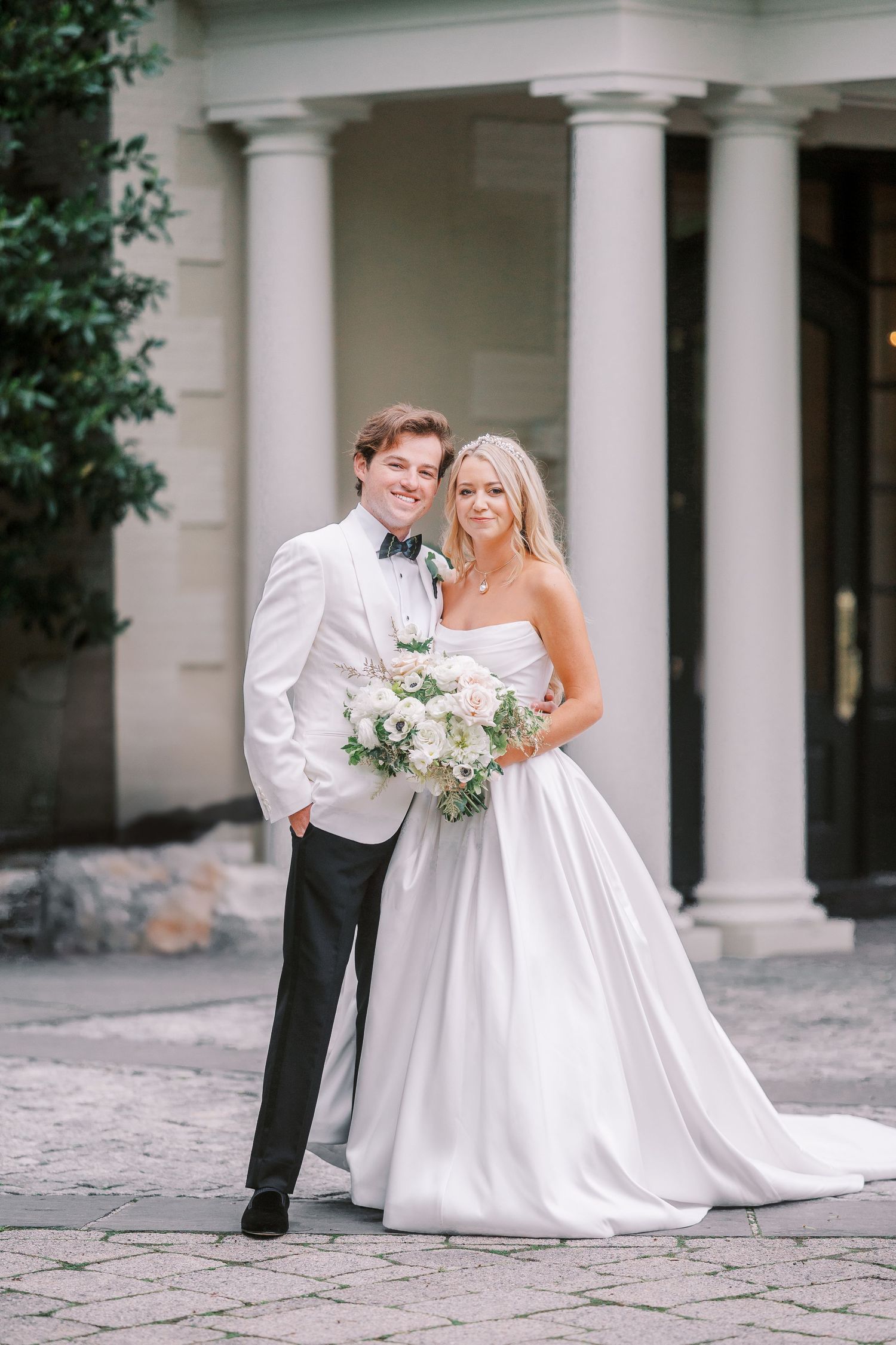 Elegant wedding couple poses together on stone patio with white columns in timeless formal attire with floral bouquet.