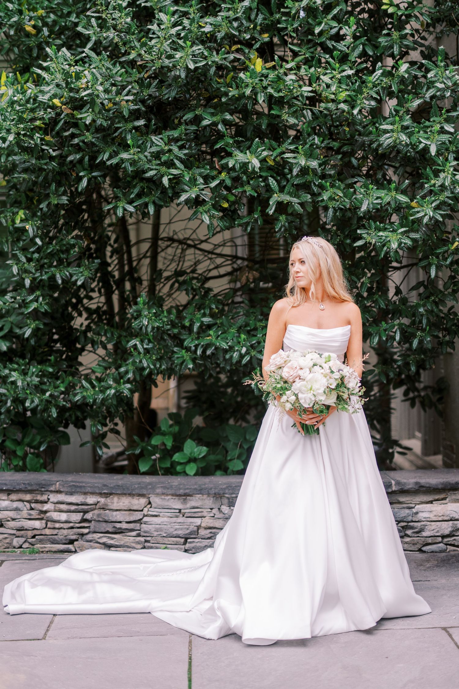 Elegant bridal portrait in a white wedding gown holding a lush bouquet against a backdrop of green foliage and stone wall.