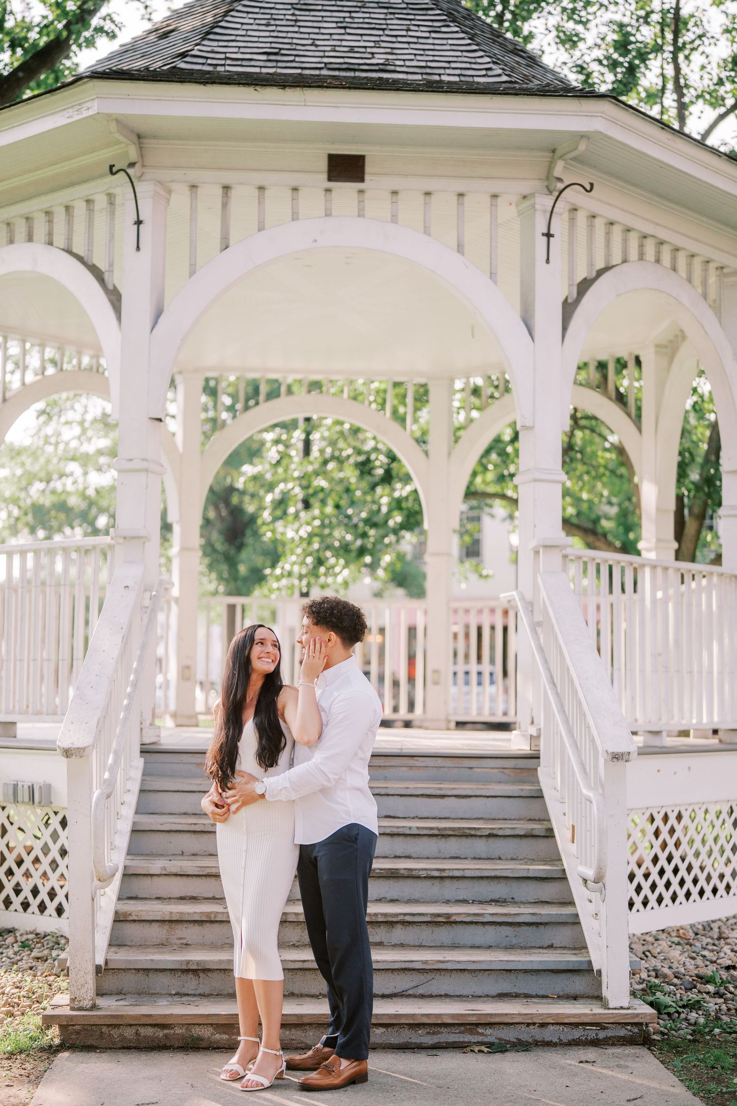 A couple shares a romantic moment on the steps of a white wooden gazebo in a lush garden setting.