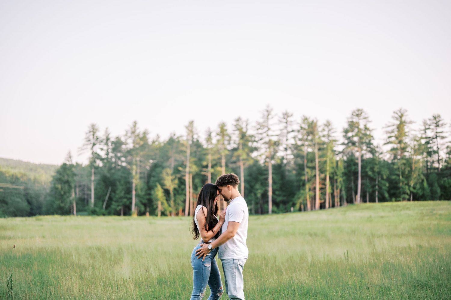 Couple embracing in beautiful meadow with tall pine trees at sunset.
