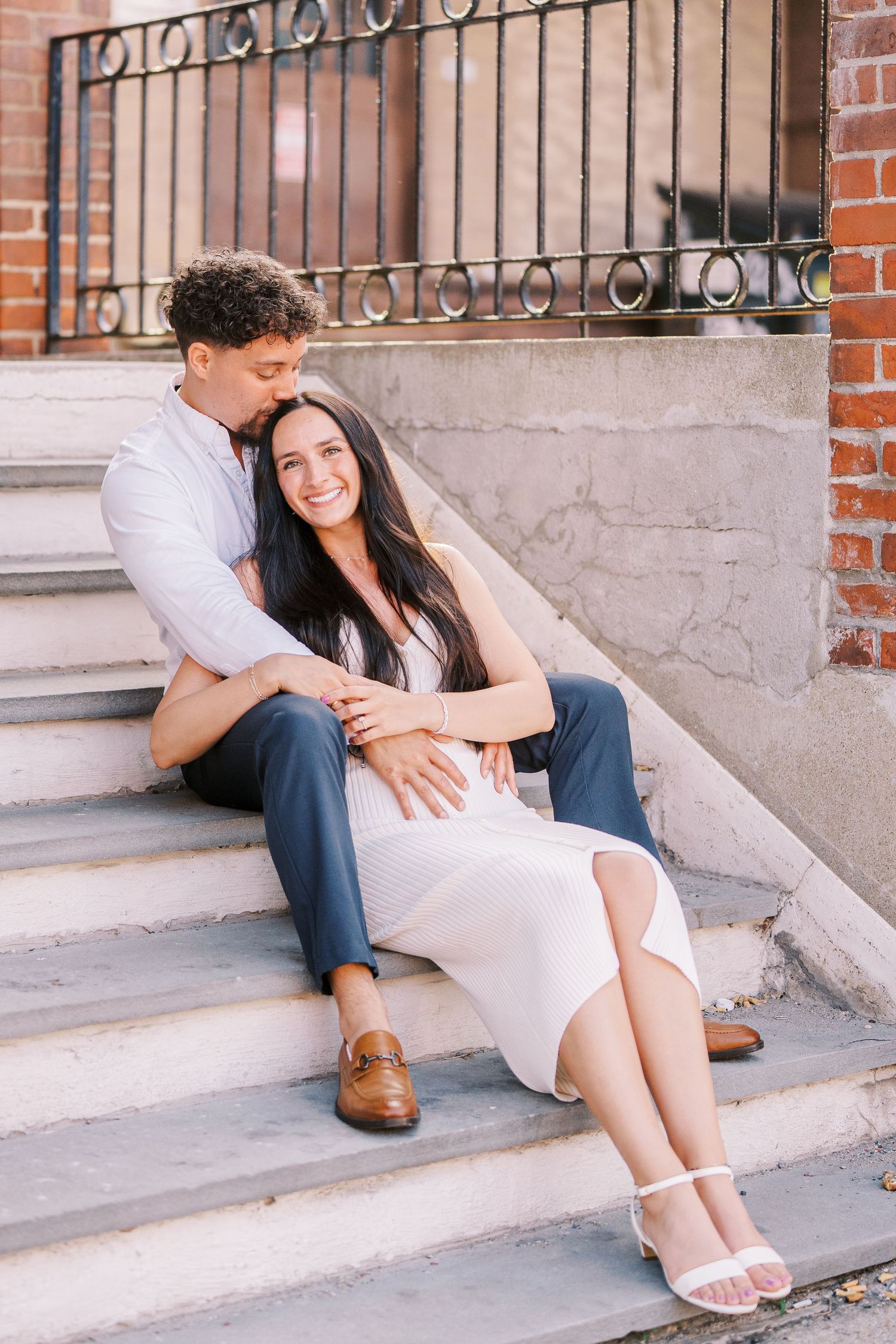 A couple sitting intimately on concrete steps in front of a brick building with decorative iron railings.
