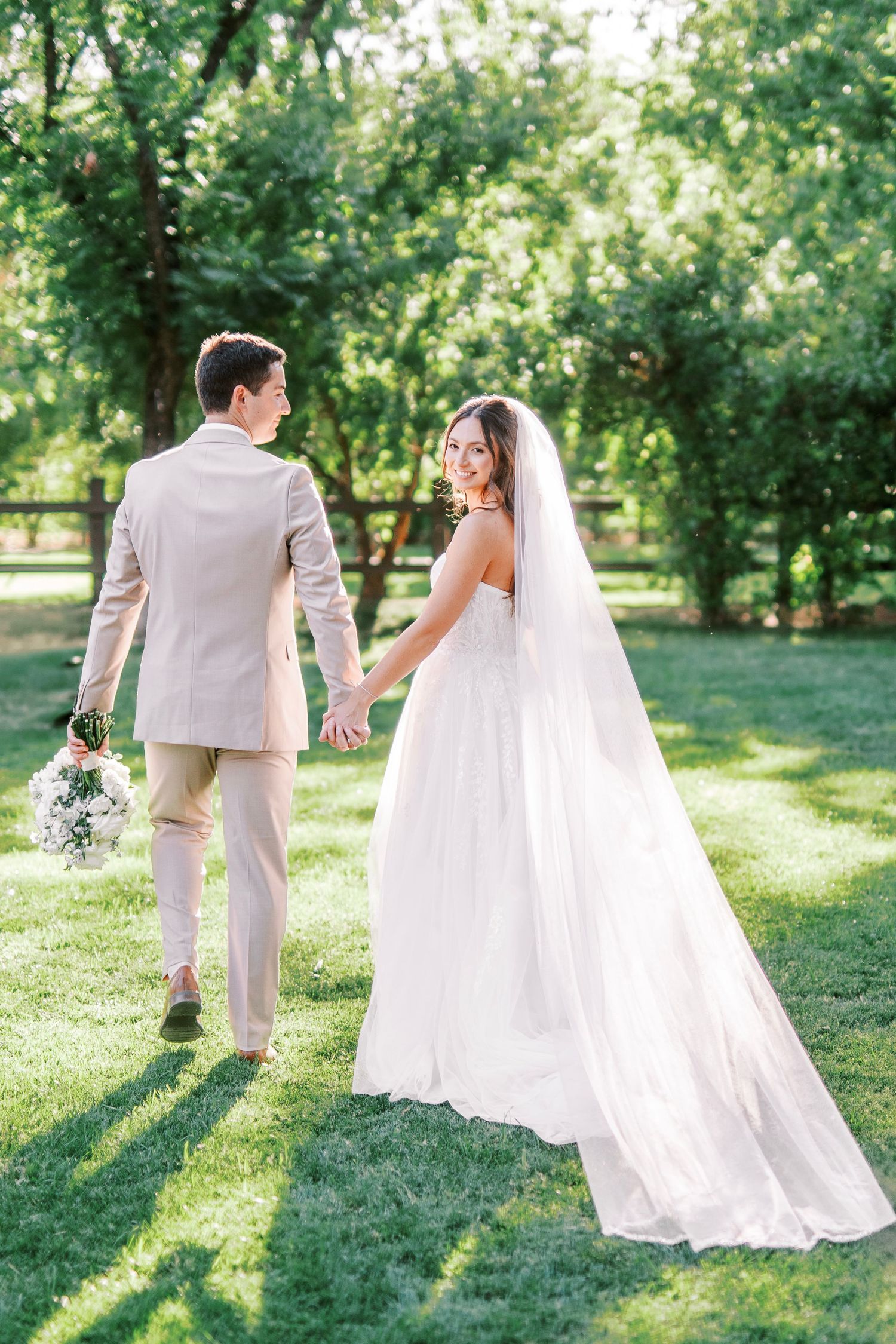 Newlyweds walk hand in hand on sunlit grass with flowing white veil and beige suit on their wedding day.