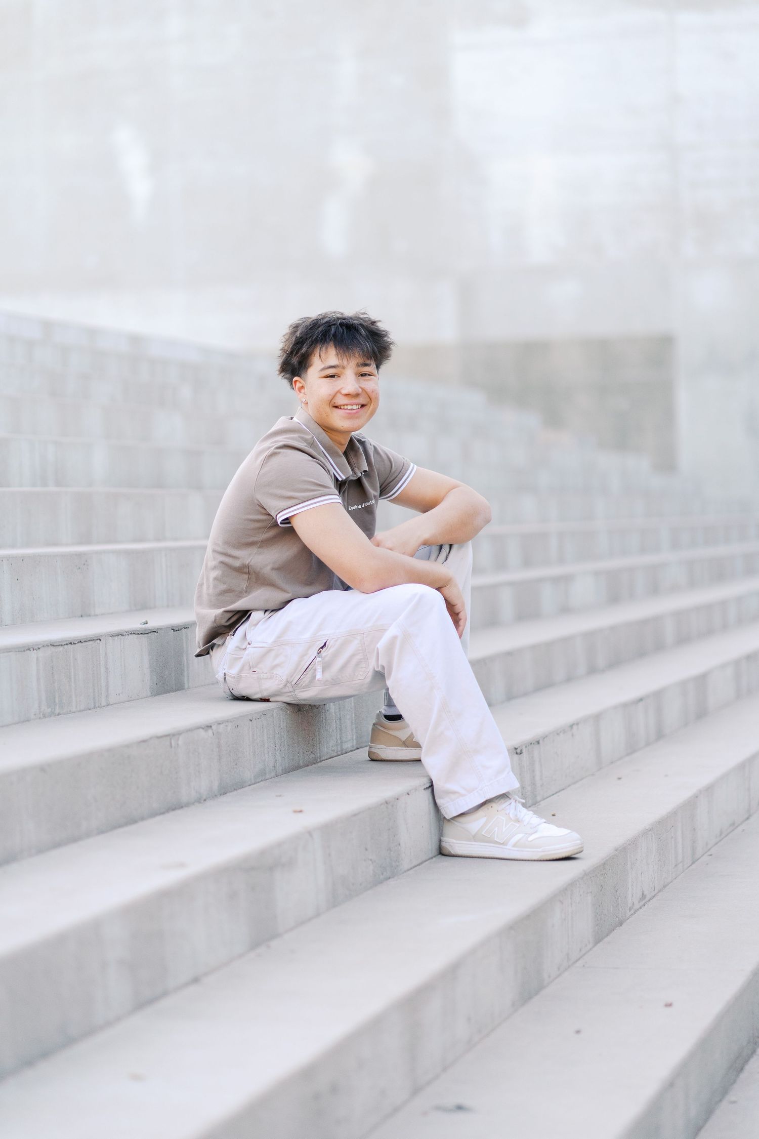 Casual portrait of someone sitting on gray concrete steps wearing beige and white against neutral background.