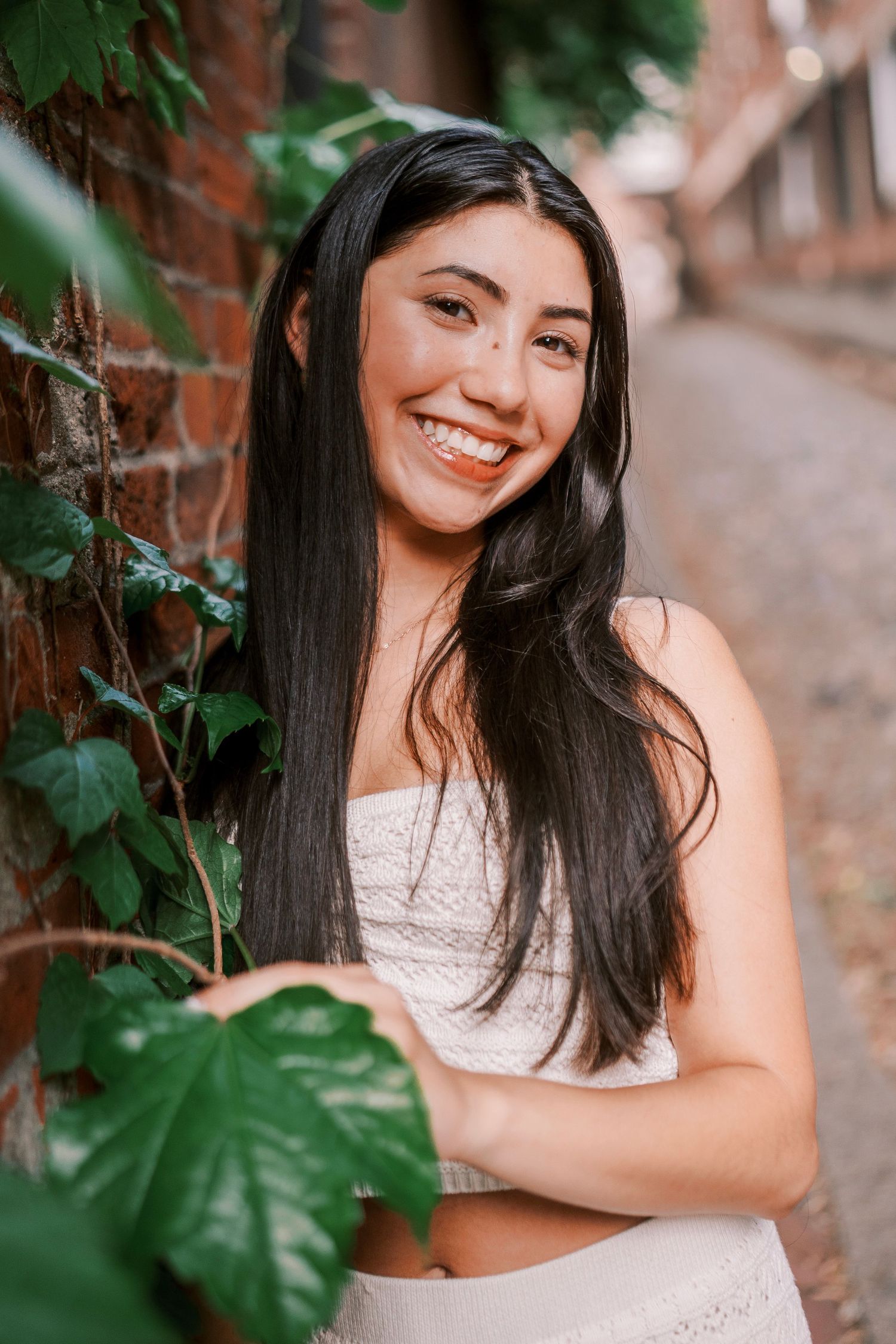 Portrait of a woman in white outfit leaning against ivy-covered brick wall while smiling warmly.