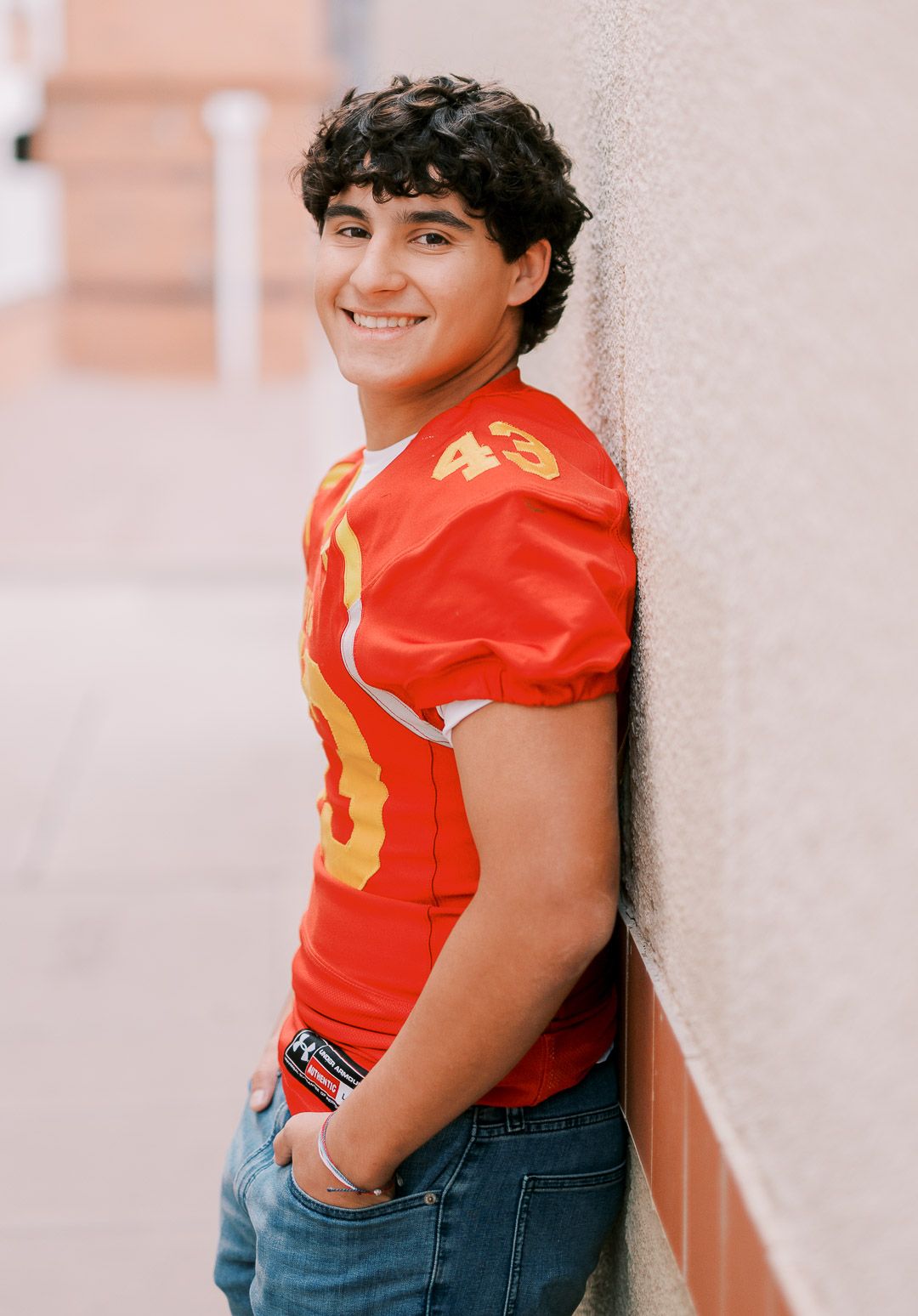A student athlete poses against a wall wearing a bright red football jersey number 43 and blue jeans.