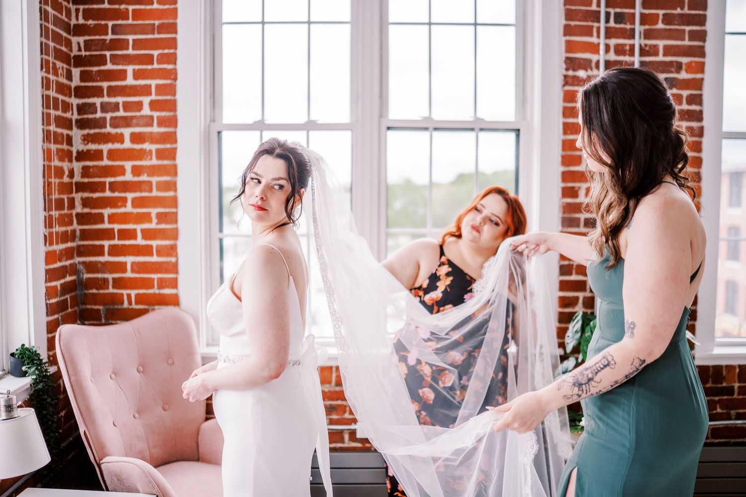 Wedding preparations in a sunlit industrial room with exposed brick walls and elegant pink accent furniture.