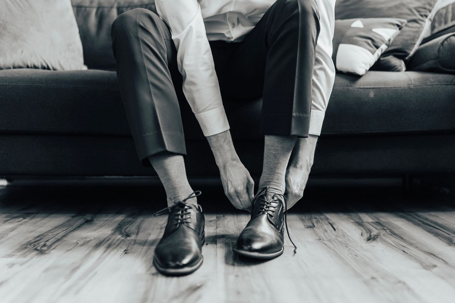 Person in formal attire sitting on dark couch tying black leather dress shoes on light wooden floor.