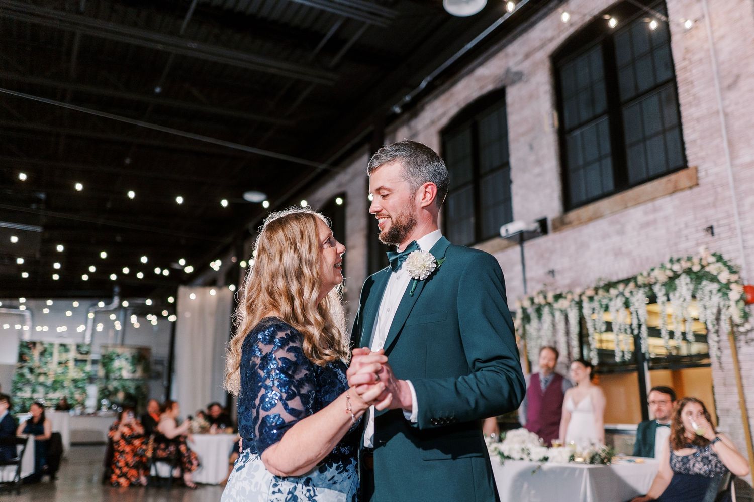 A couple shares an intimate dance moment at an indoor reception venue decorated with string lights and greenery.