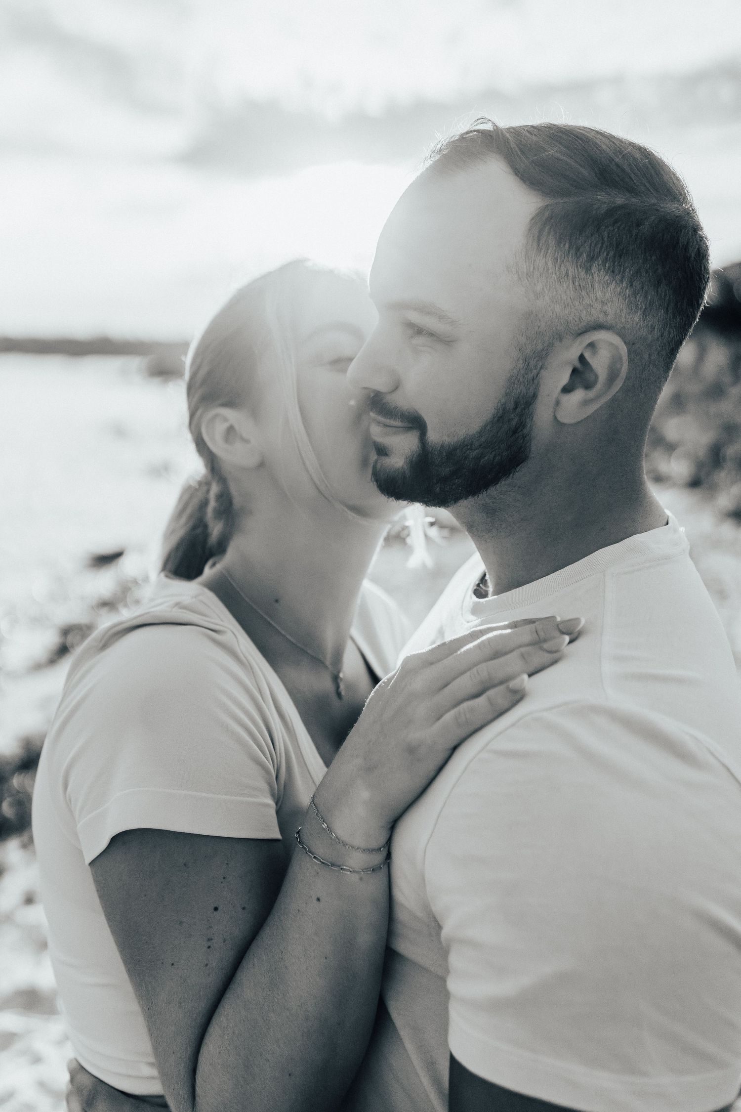 Intimate black and white portrait of couple embracing outdoors with sunlit background.