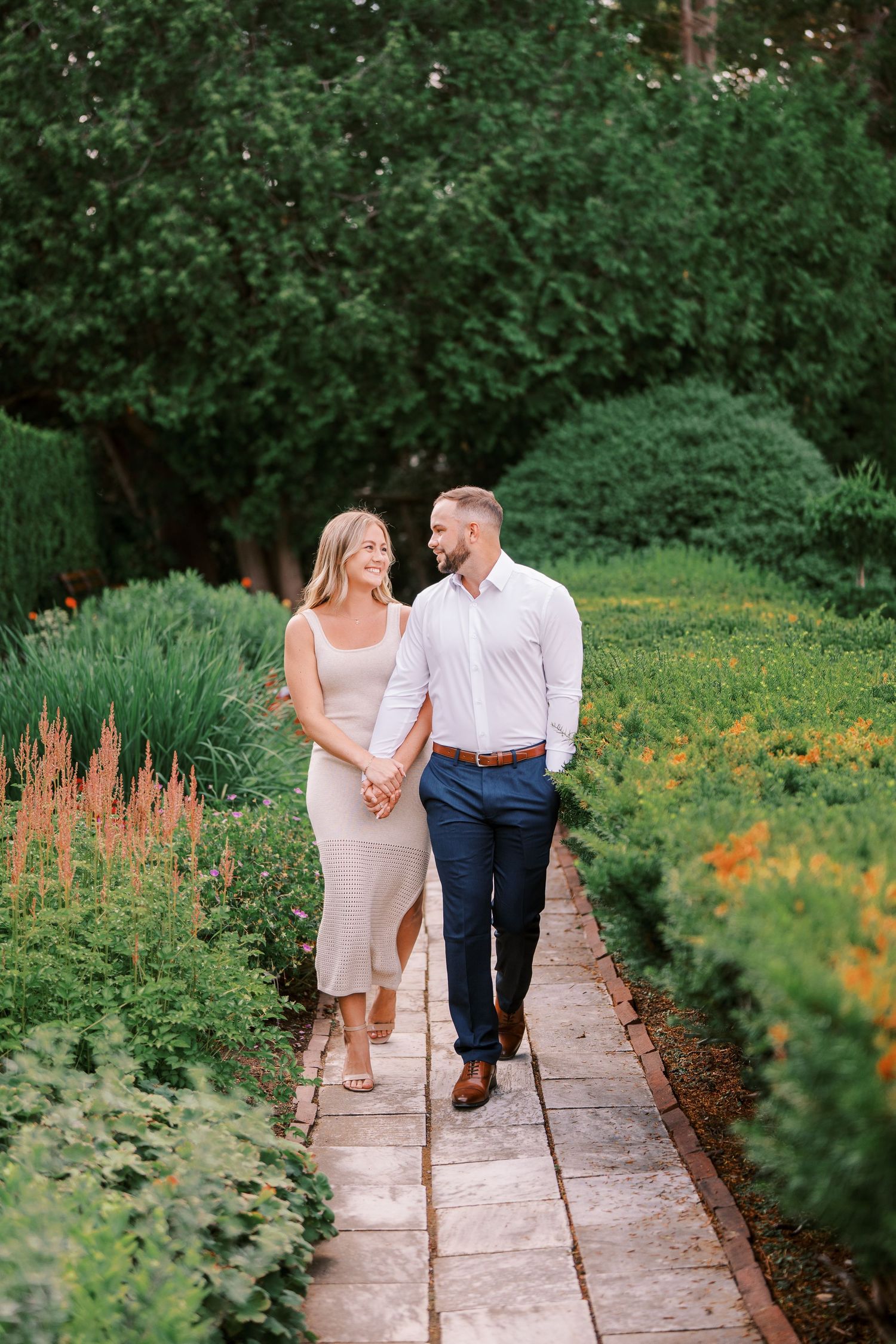 Couple walking hand in hand along garden path with lush greenery and flowers.