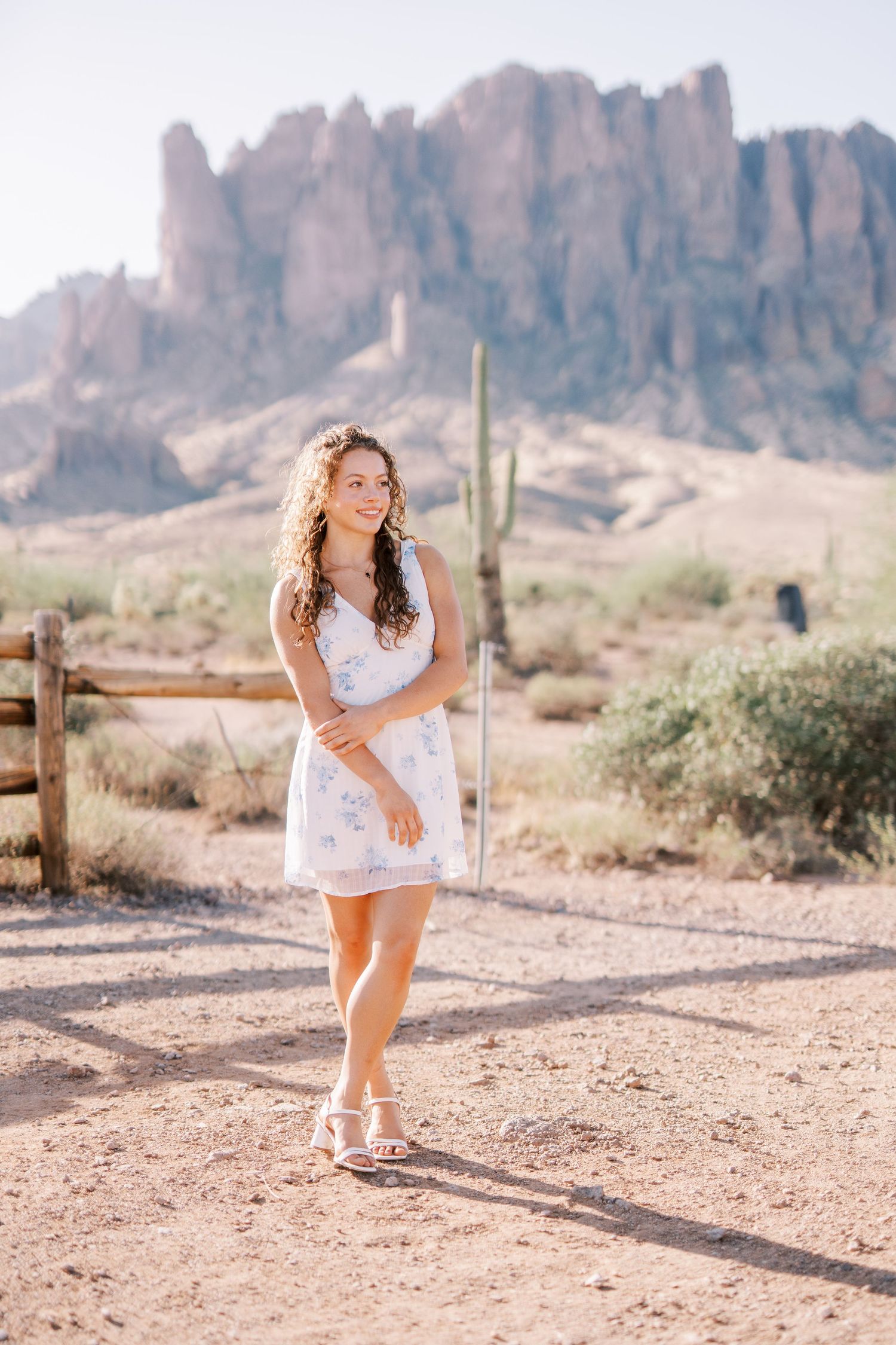 A woman in a white dress poses in the desert with rugged mountains and a saguaro cactus in the background.