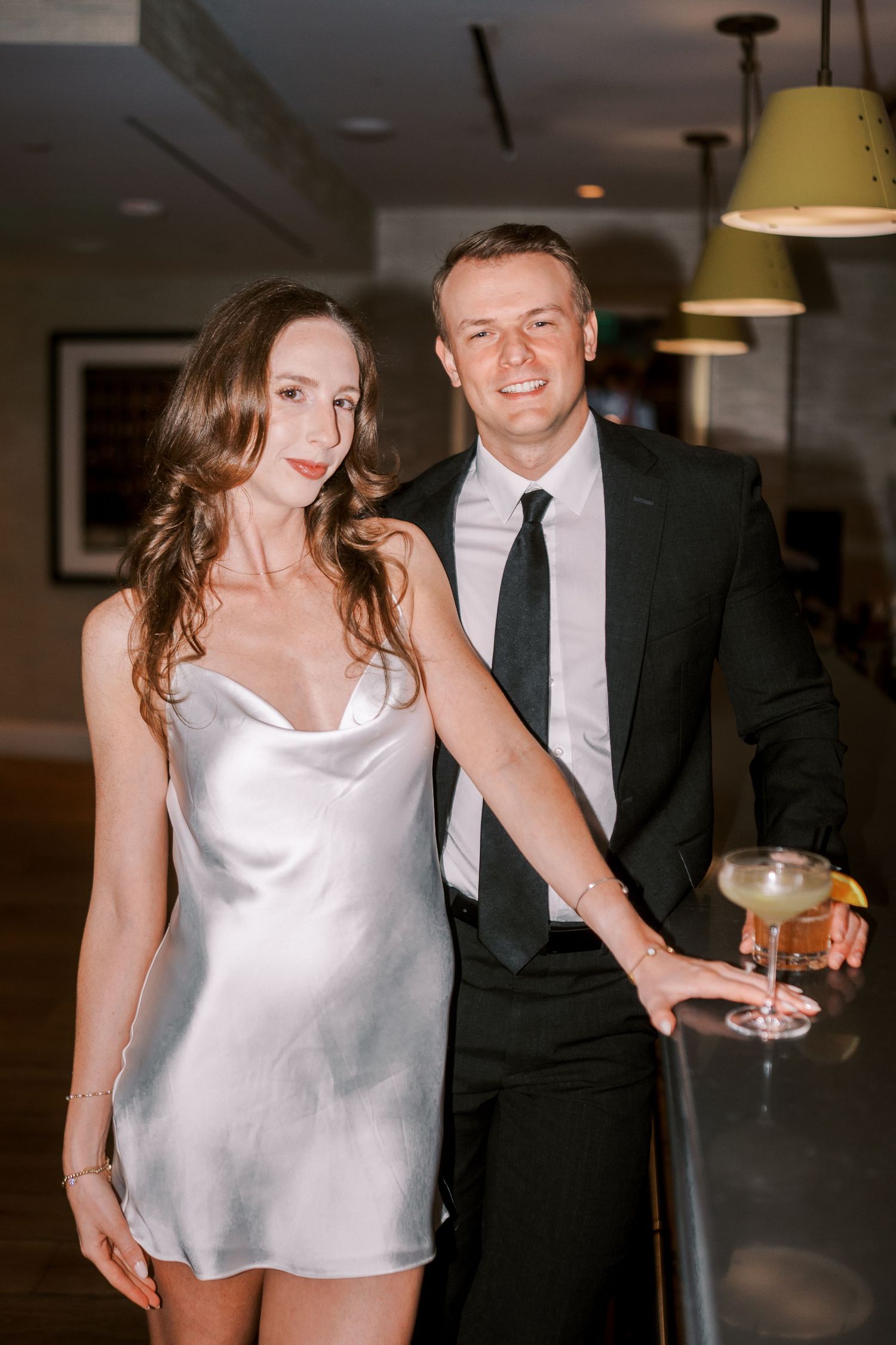 Couple at formal event with cocktails, woman in silk slip dress and man in black suit and tie posing together at bar.