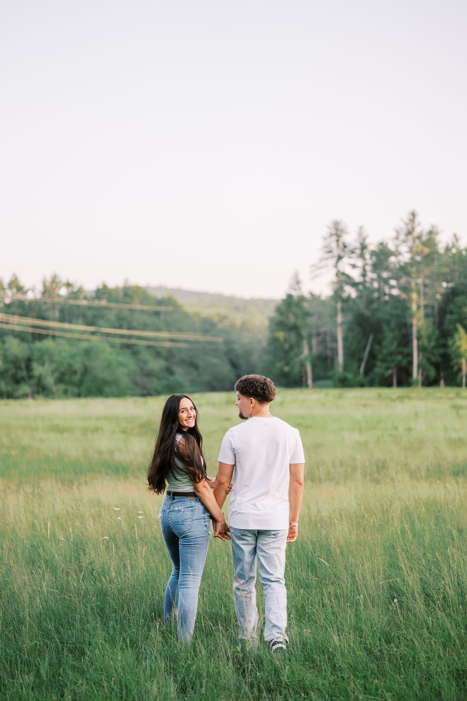 A romantic couple holding hands while walking through a serene meadow with trees and hills in the background.