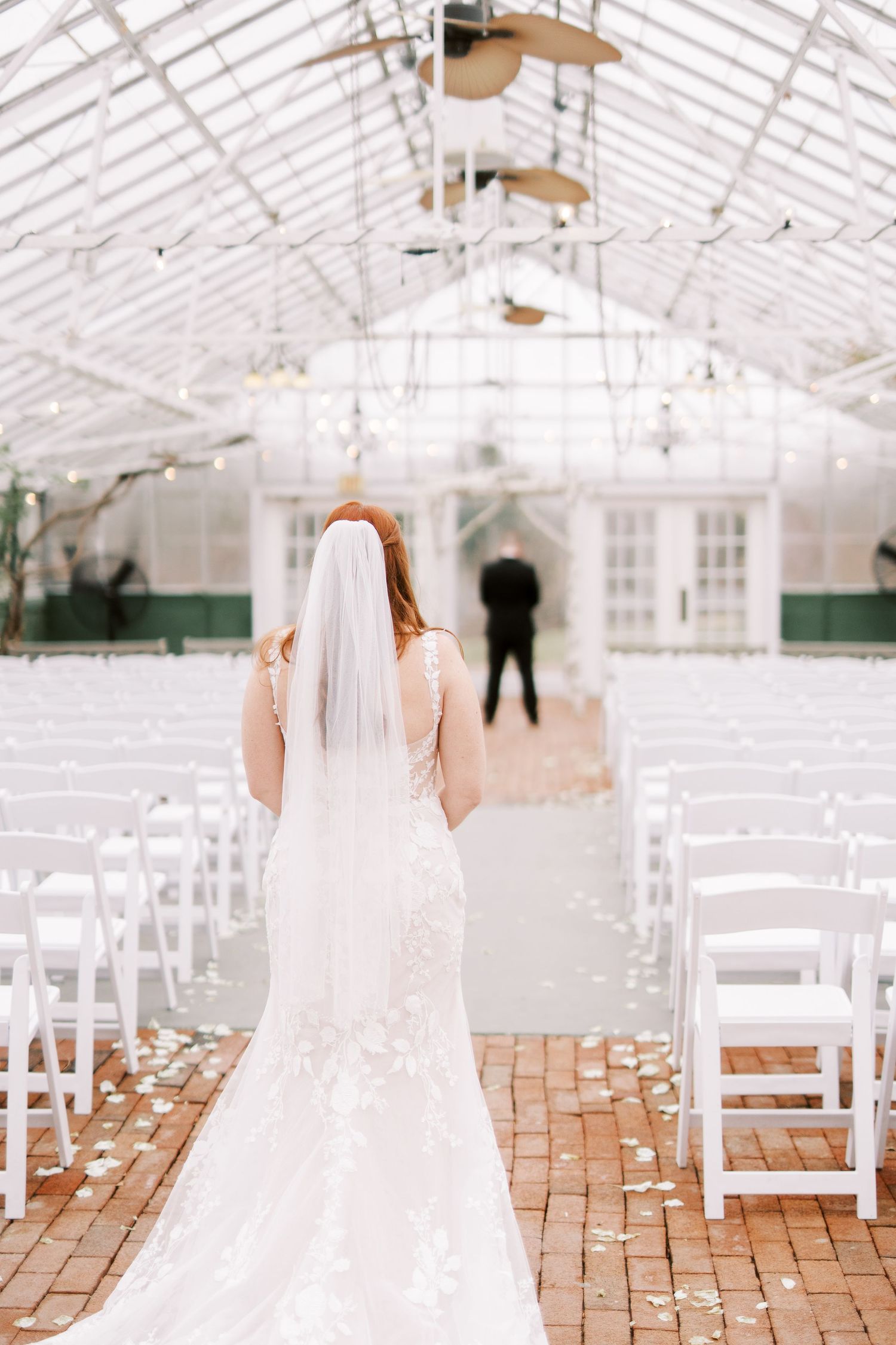 A bridal ceremony setup in a bright greenhouse with white chairs and brick aisle.