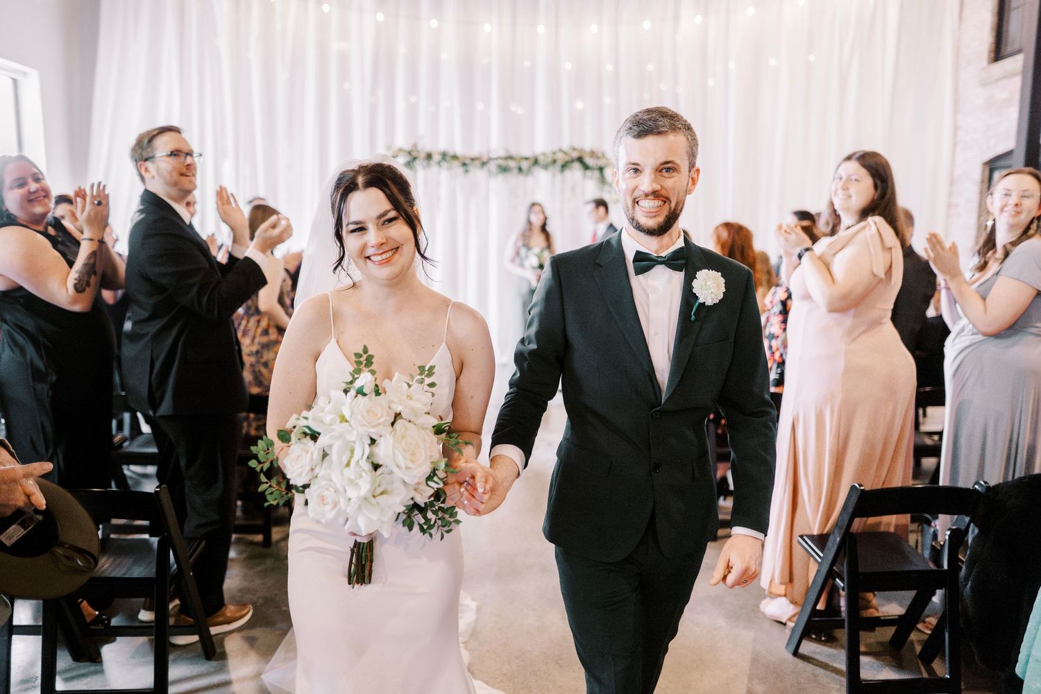 A wedding couple walks down the aisle while guests clap and celebrate in a white decorated ceremony space.