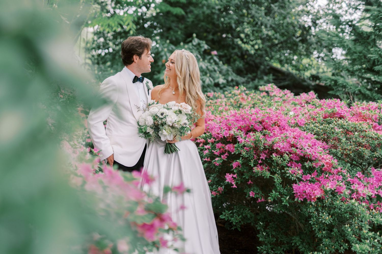 Two people hold hands and pose together in a lush garden with bright pink flowering bushes blooming in the background.