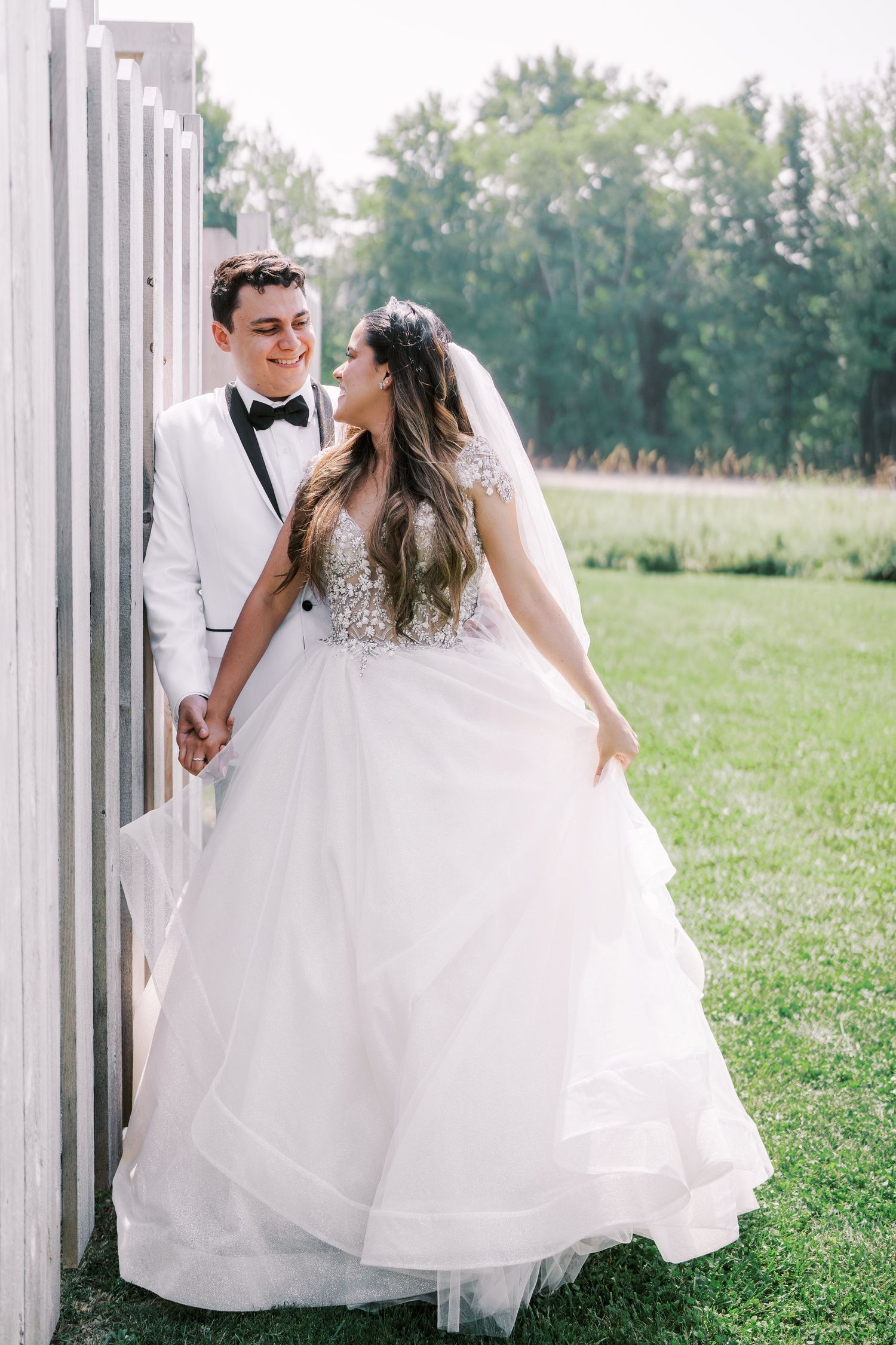 A bride in a flowing white wedding gown spins and dances joyfully in a grassy outdoor setting.