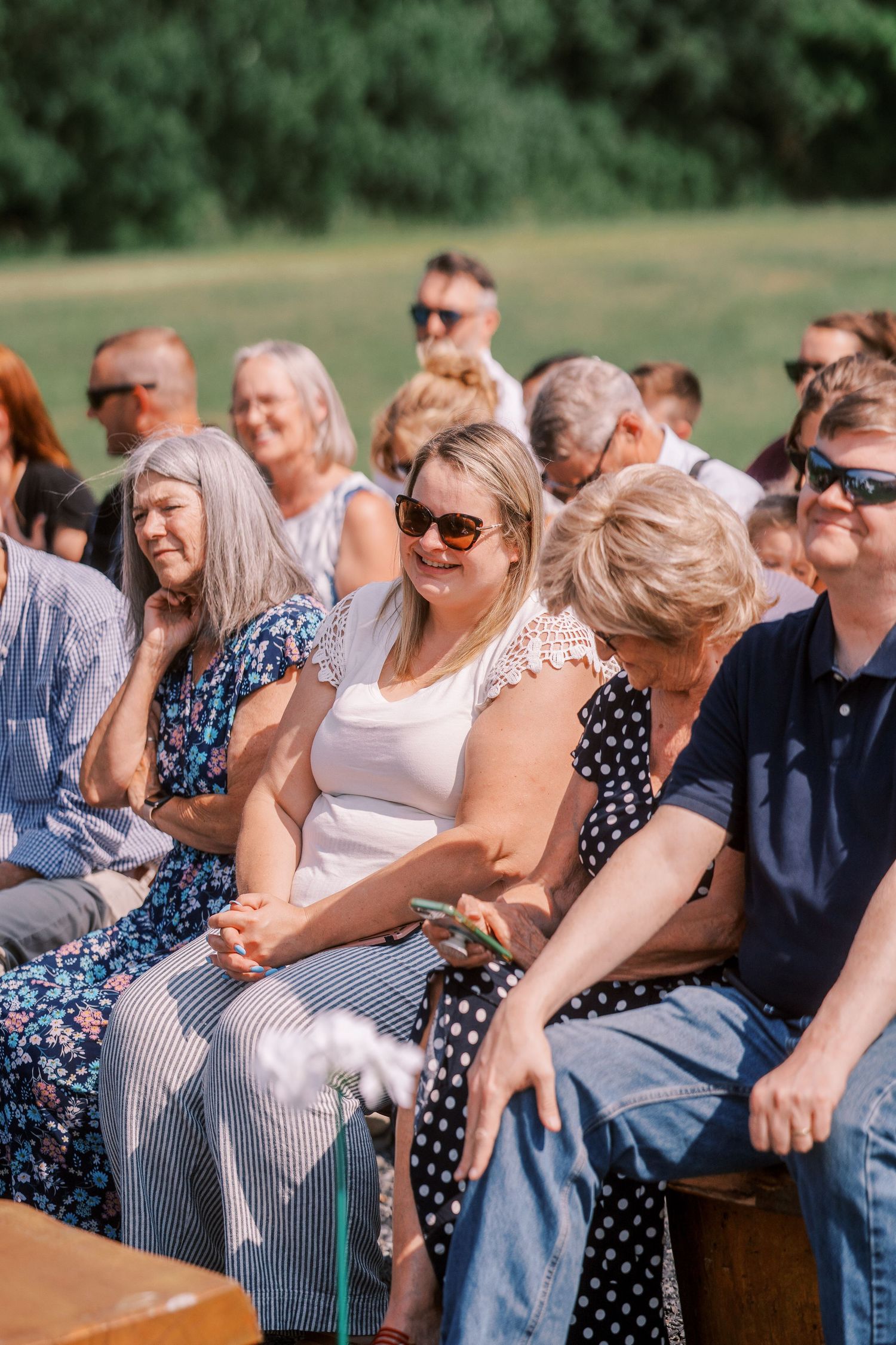 Wedding guests smile and laugh while seated at an outdoor ceremony on wooden benches in a garden setting.