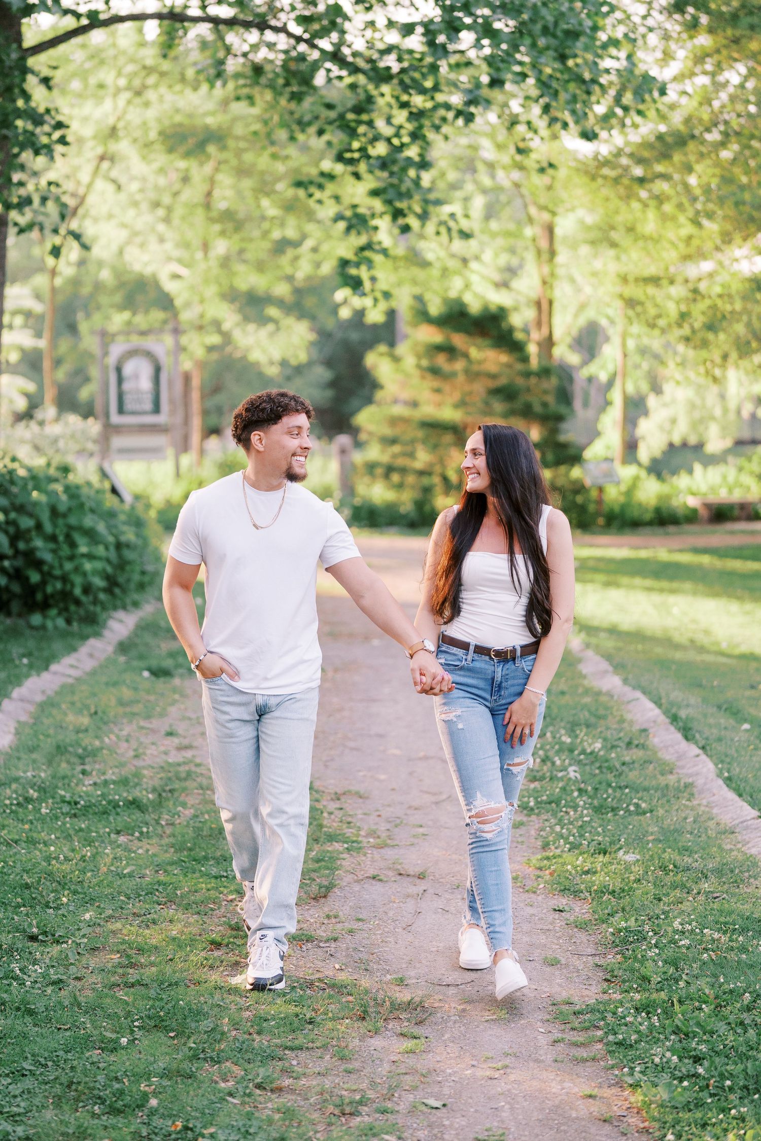 Young couple walking together and holding hands on a garden path surrounded by green trees and foliage.