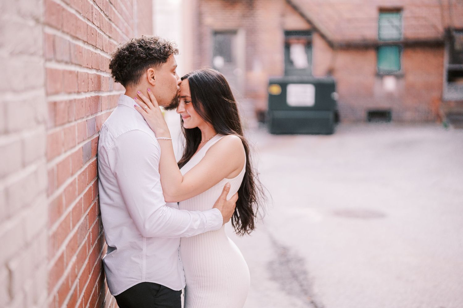 A couple shares an intimate moment against a brick wall in an urban setting.