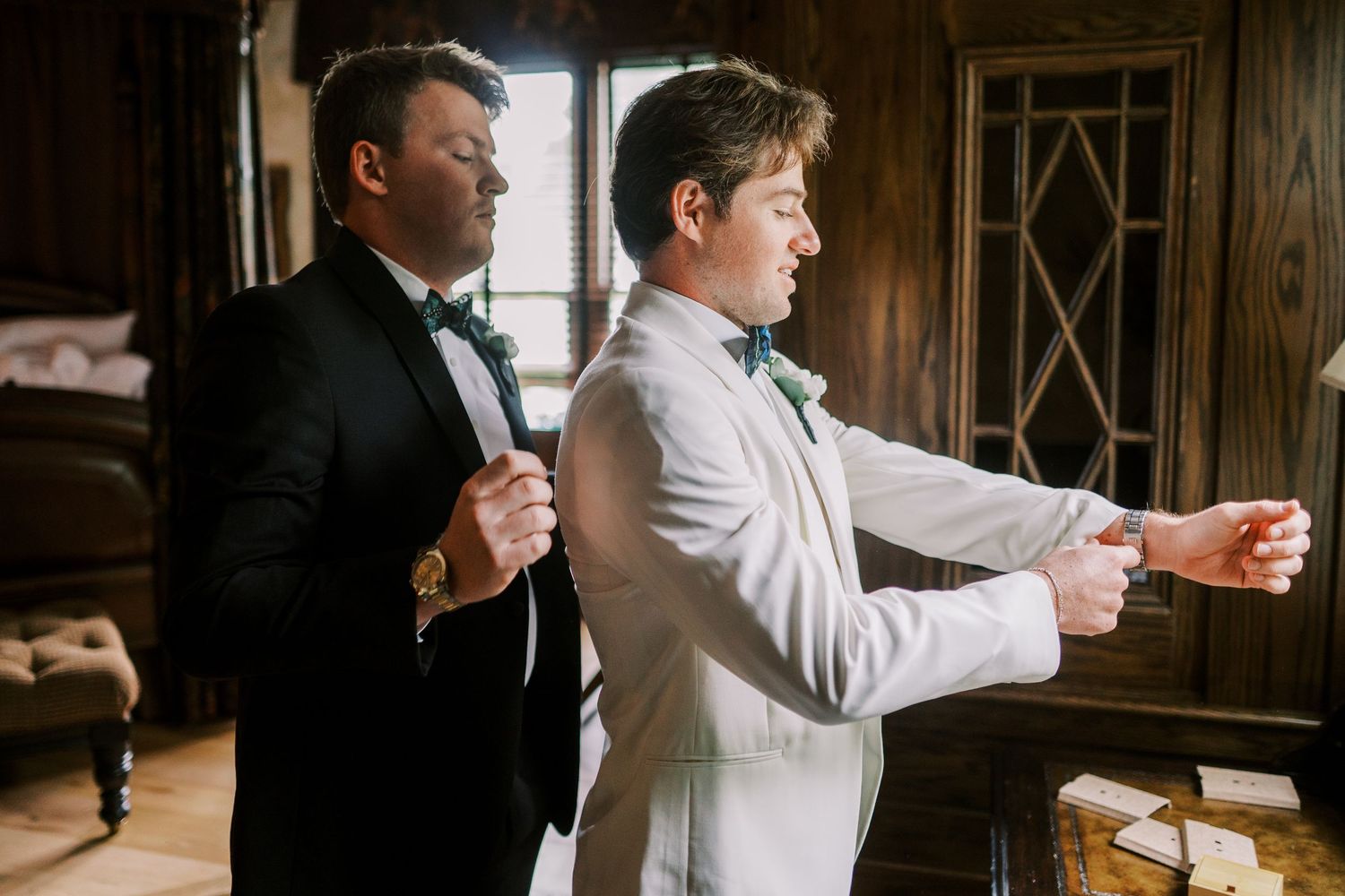 Two people in formalwear stand together, one helping the other adjust their outfit in an elegant interior setting.