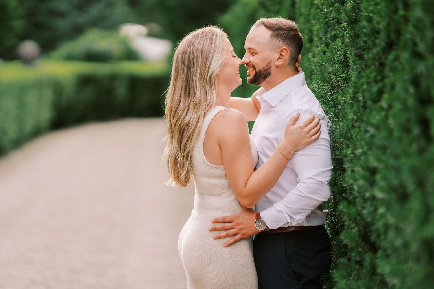 A couple embraces romantically in a garden pathway with lush green hedges surrounding them.