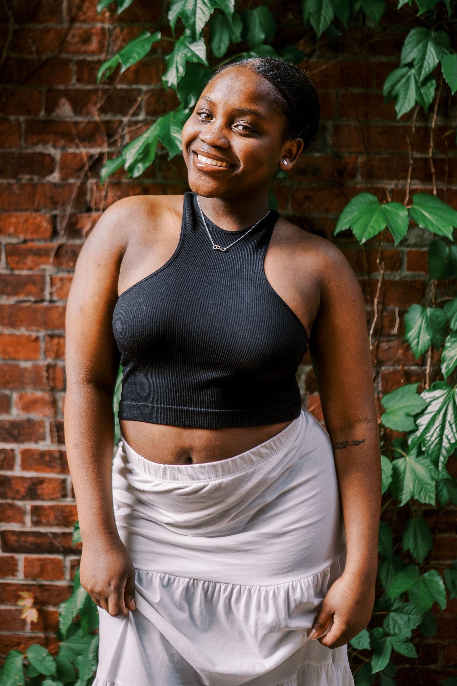 Portrait against brick wall wearing black crop top and light flowy skirt with green leaves in background.