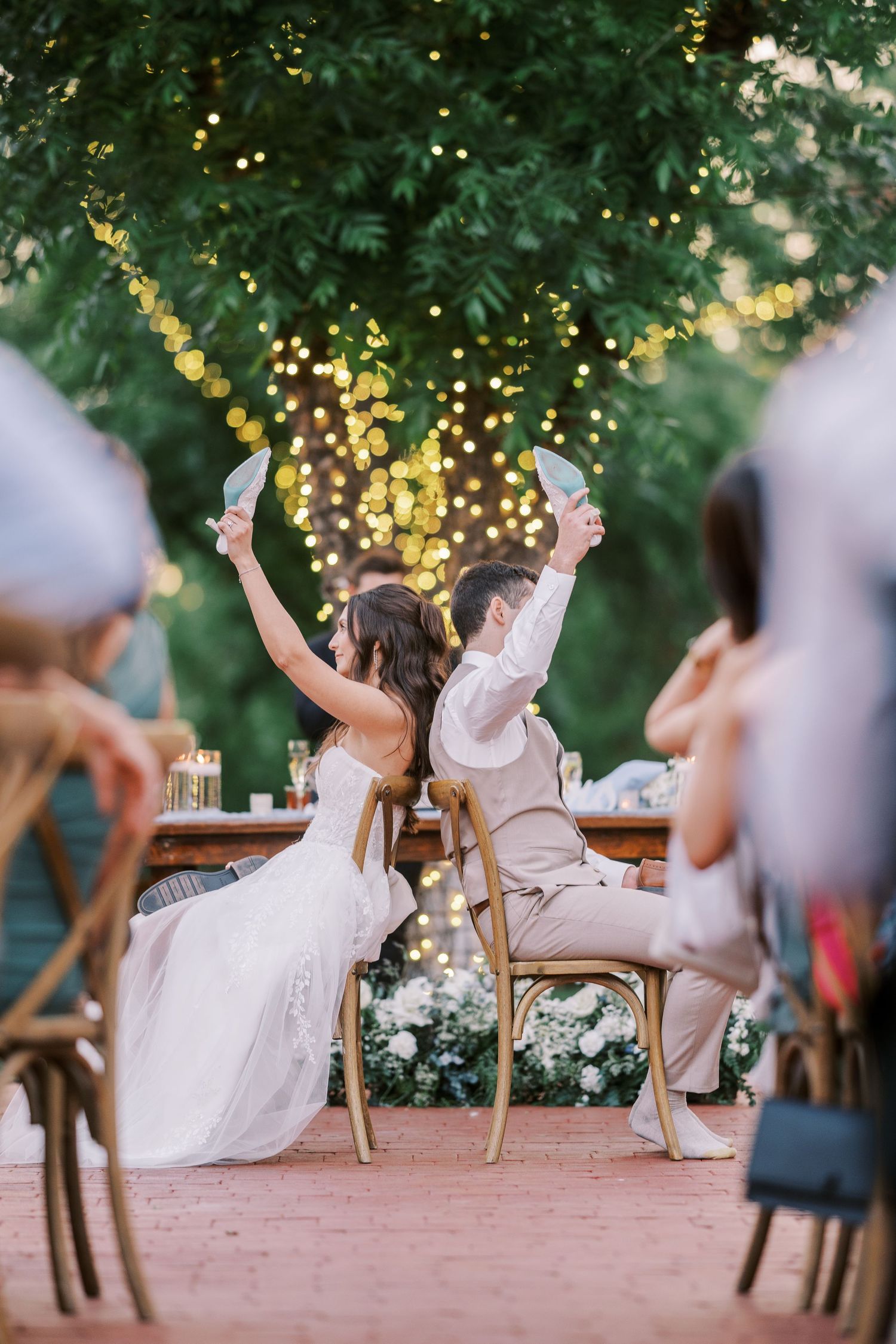 Wedding guests celebrating joyfully in chairs at outdoor garden reception illuminated by string lights at dusk.