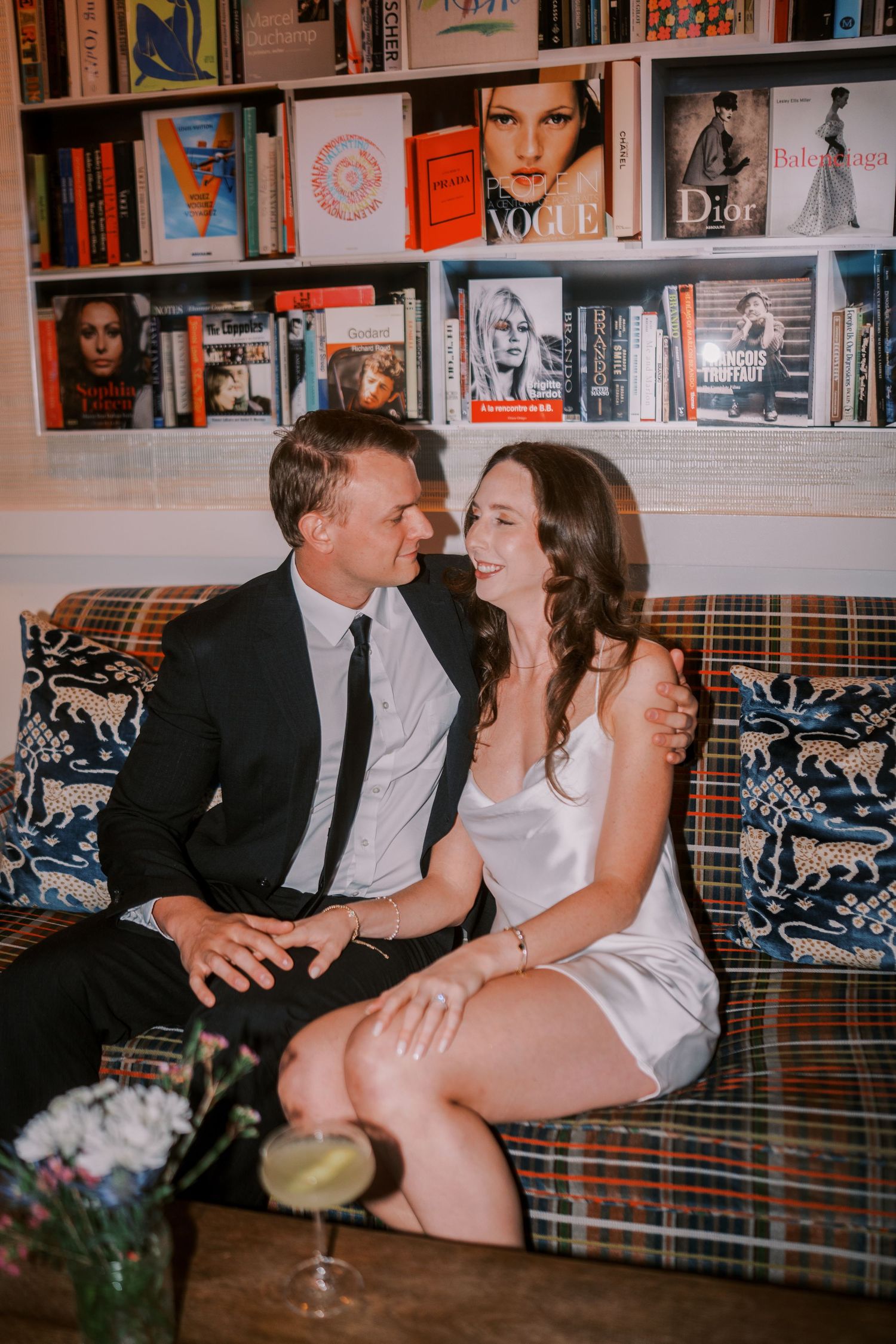 A couple shares an intimate moment on a couch with bookshelves displaying fashion magazines and portraits in the background.