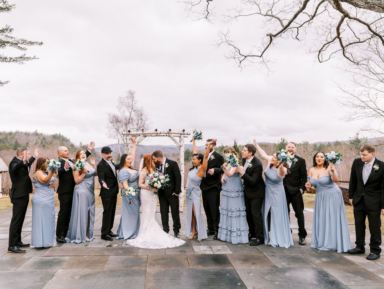 A wedding party lined up outdoors wearing blue dresses and dark suits against an overcast sky.