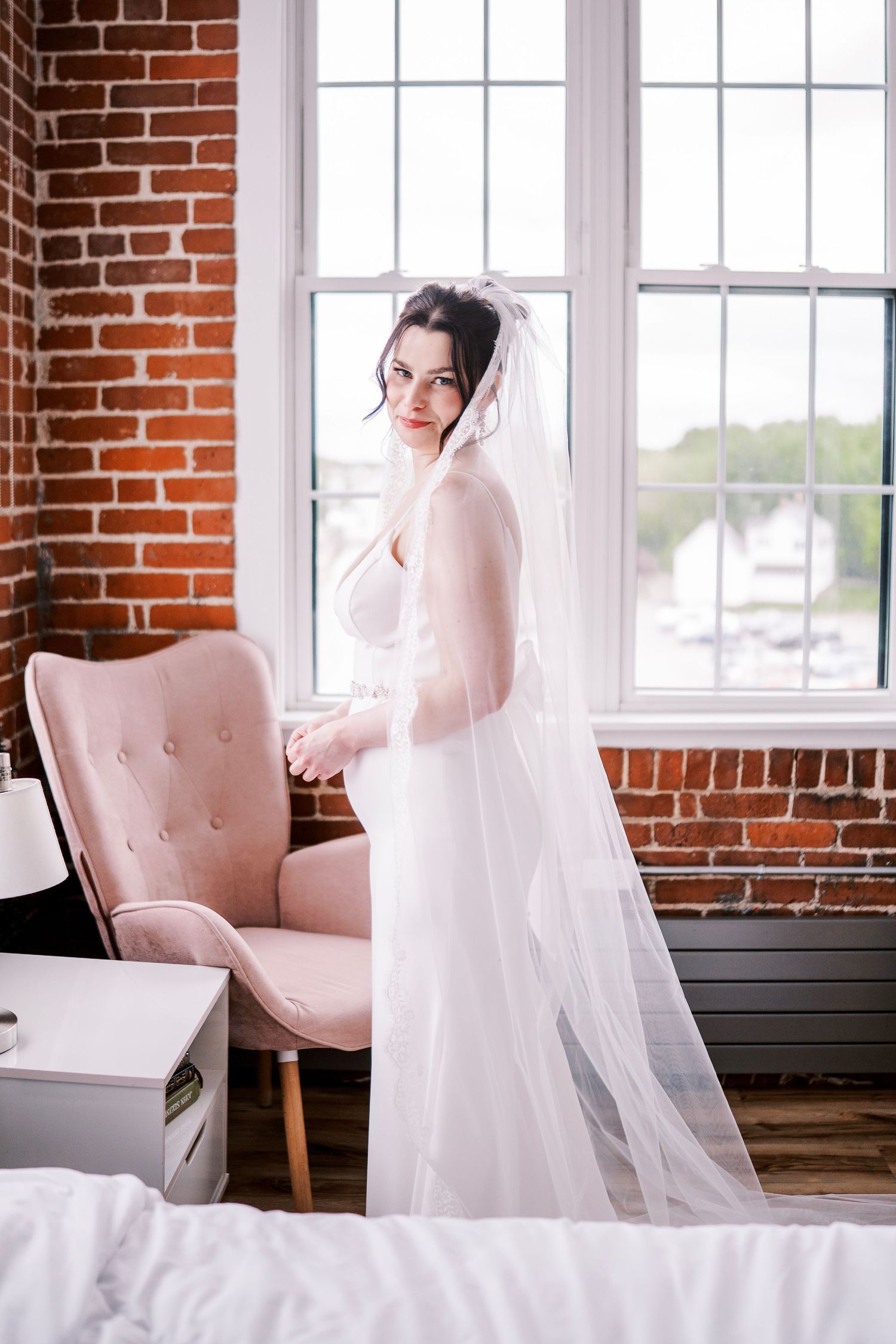 A bride in a long veil poses elegantly next to a pink accent chair by large windows with exposed brick walls.